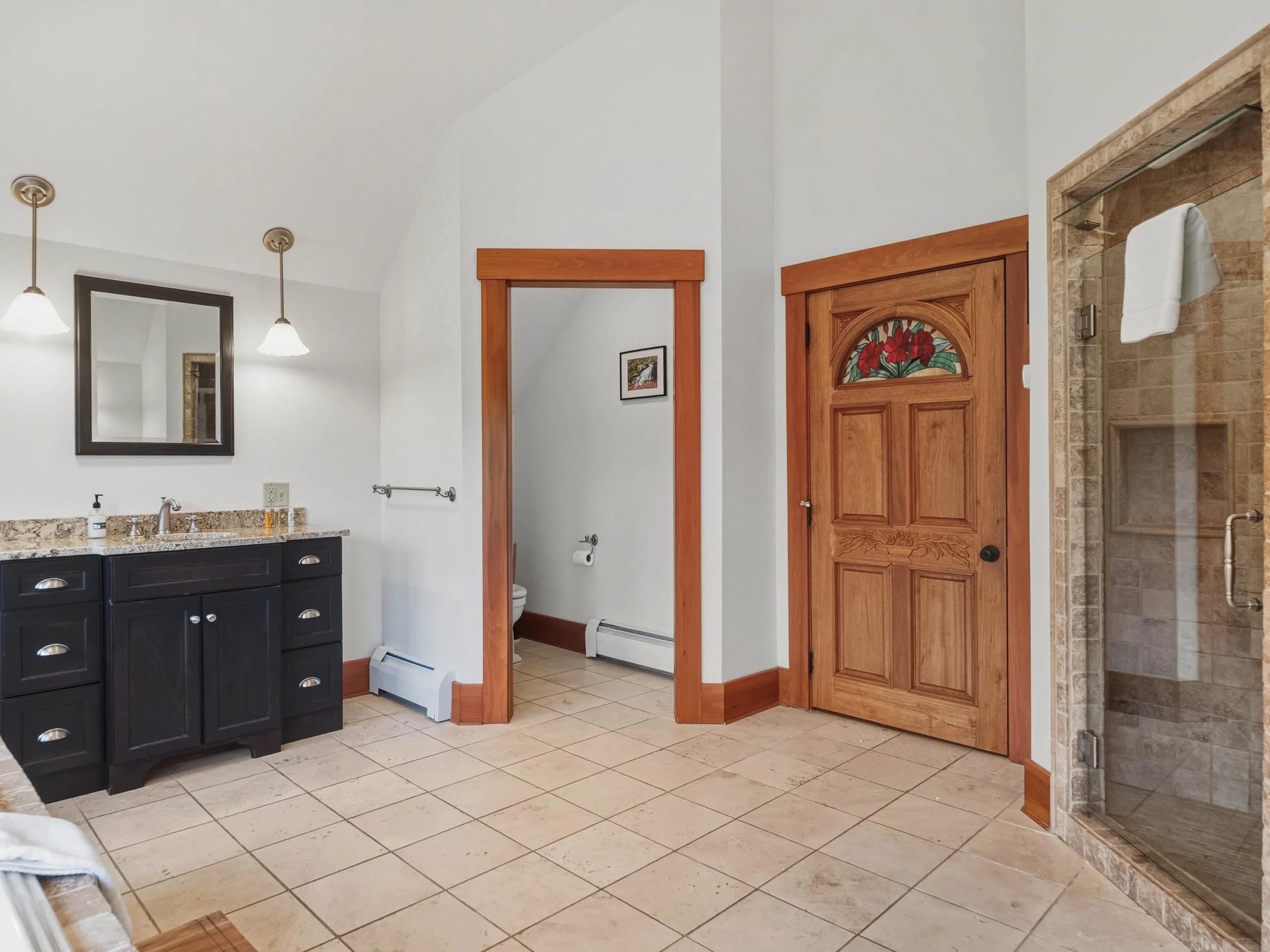 Bathroom with a black vanity, granite countertop, mirror, two hanging lights, a mirror, a small picture, a toilet, a closed wooden door with stained glass, a glass shower with beige tiling, and tiled flooring.