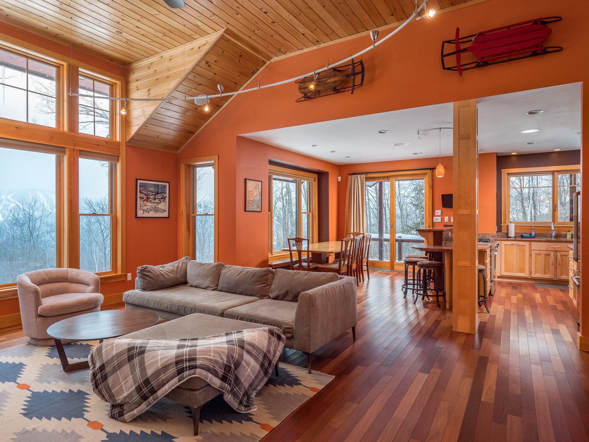 Modern living room and dining area with orange walls, wooden ceiling, large windows, gray sofa, armchair, coffee table, dining table with chairs, kitchen area, wooden floors, and decor items like sleds on the wall.