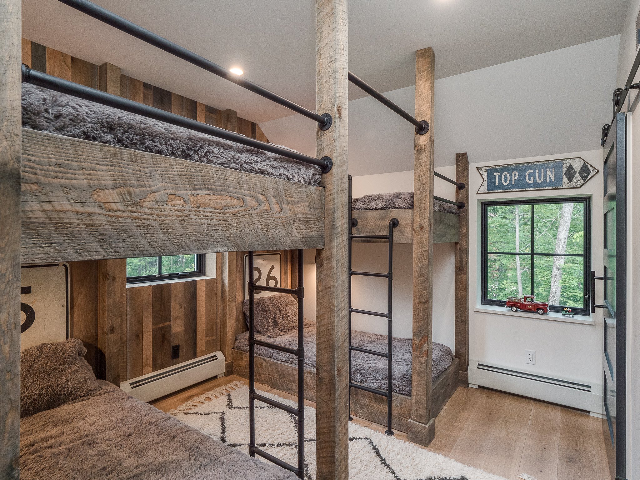 Rustic bedroom with wooden bunk beds, gray bedding, "Top Gun" sign, rug, and window view of trees.