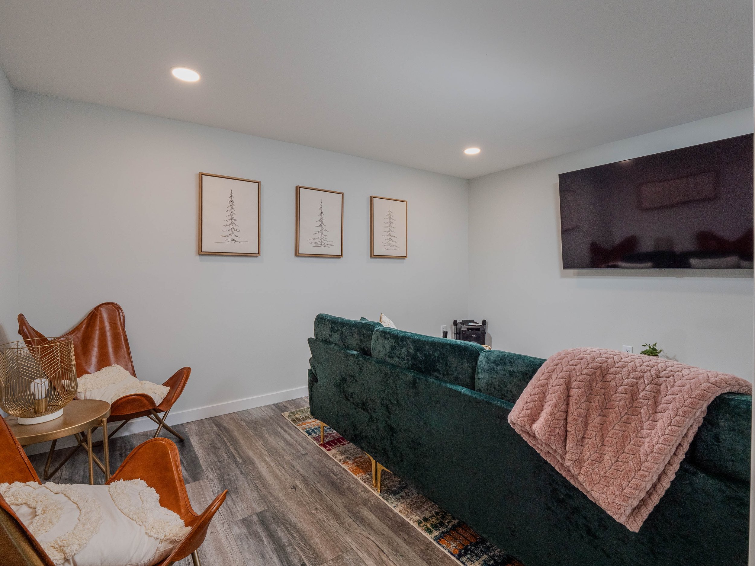 Living room with a green velvet sofa, pink textured blanket, two leather chairs, a wall-mounted TV, and framed minimalist tree art on a light blue wall.