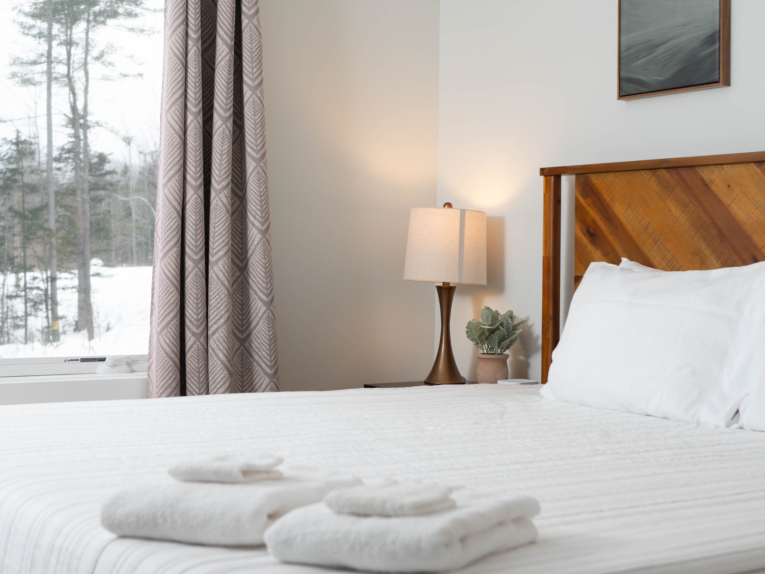 Inside a bedroom with a bed, white pillows, and folded towels on the bed, a wooden headboard, a bedside lamp, a potted plant, a window with patterned curtains showing a winter landscape outside, and a framed painting on the wall.