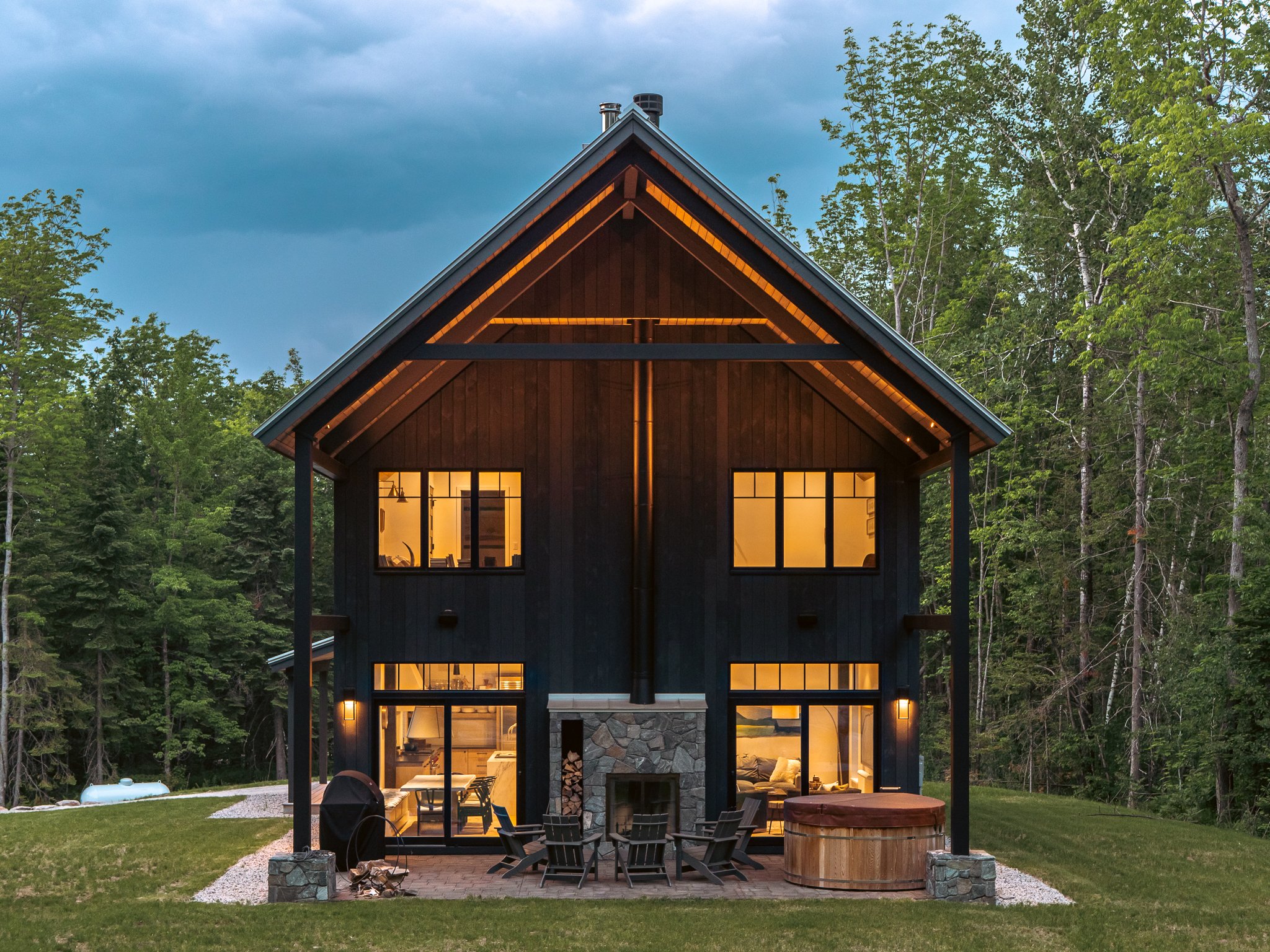 Modern black wooden house with large windows, outdoor seating, and hot tub surrounded by trees.