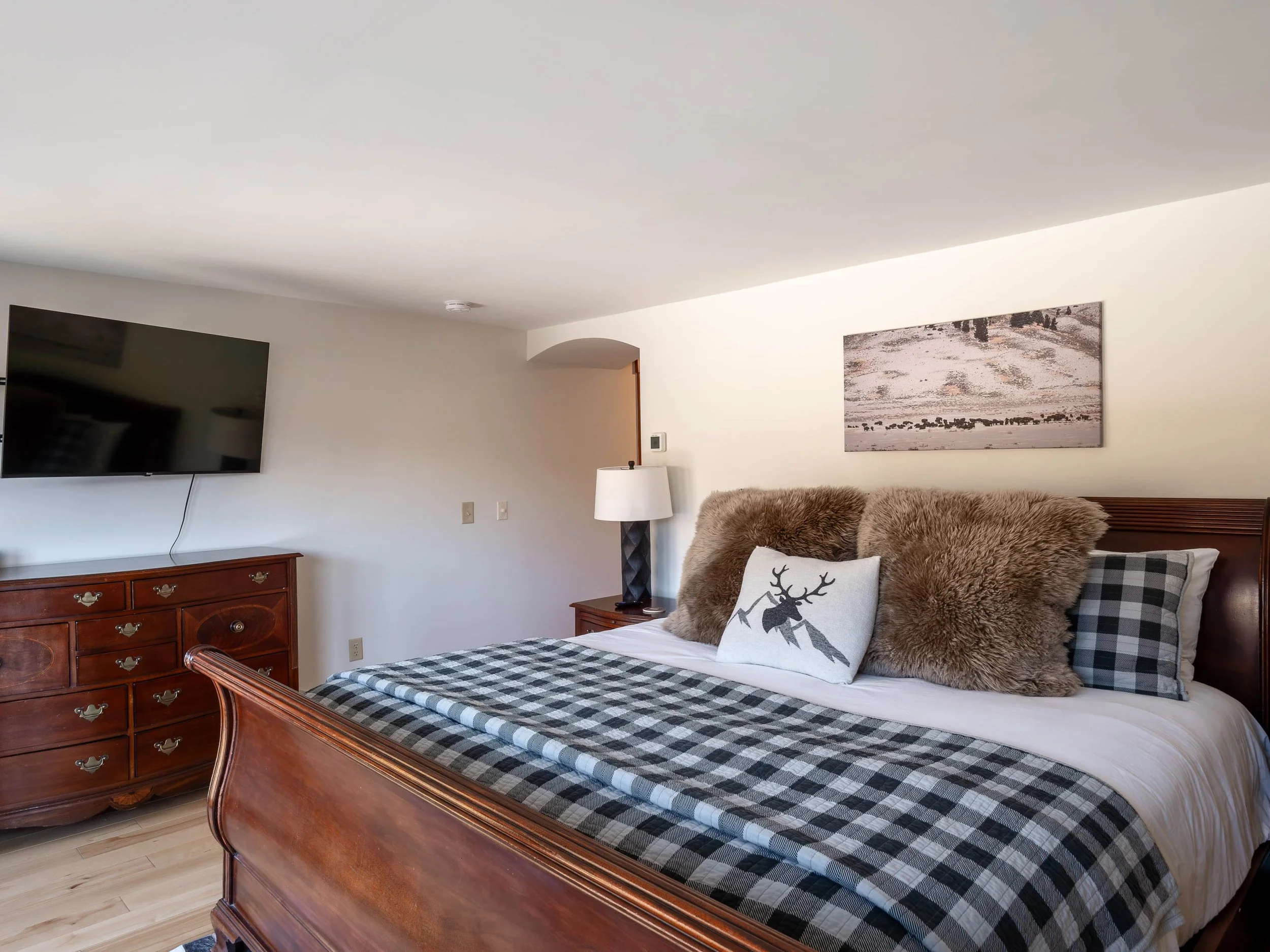 A bedroom with a wooden bed frame, plaid bedding, fur pillows, and a decorative pillow with a deer head. There is a wooden dresser with a television above it, a nightstand with a lamp, and a landscape painting on the wall.