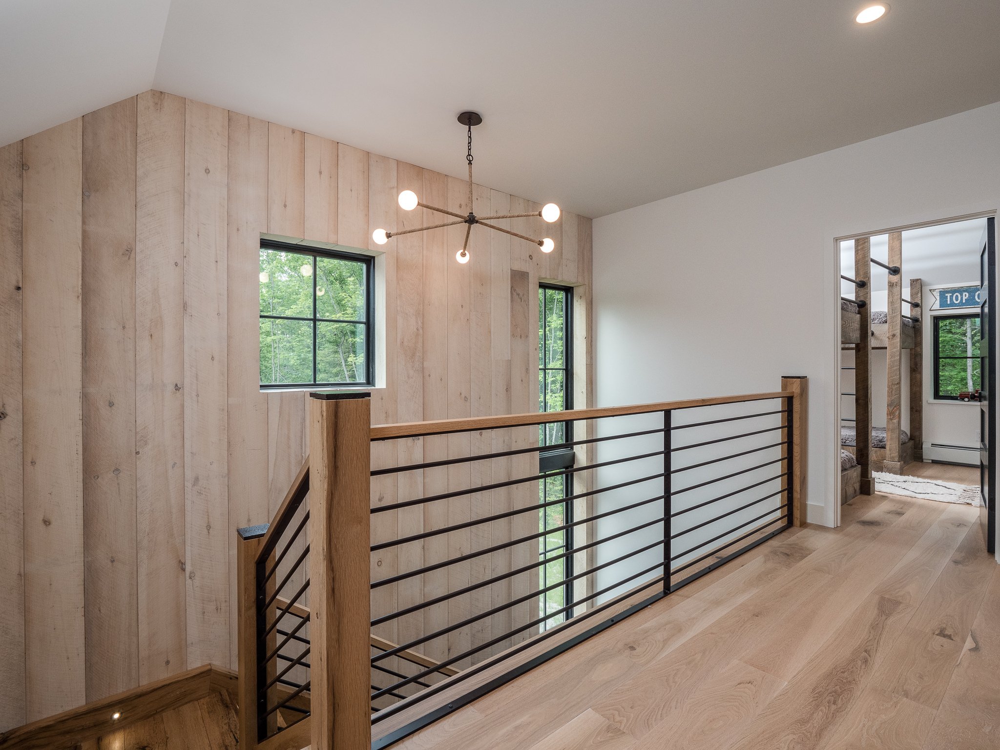 Modern hallway with wooden floors and walls, featuring a metal railing. A chandelier hangs from the ceiling. A doorway leads to a room with bunk beds.