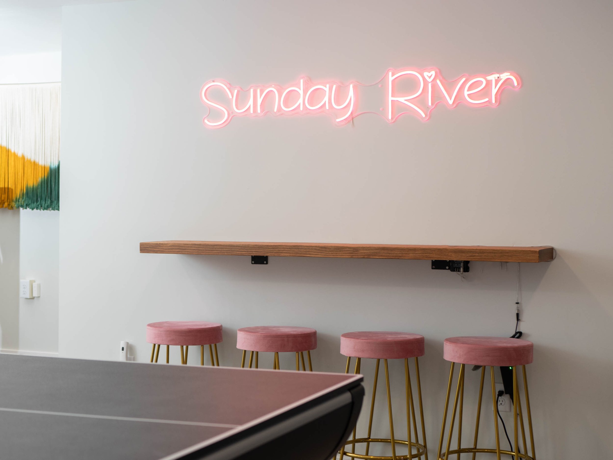 Interior space with a neon pink sign that reads 'Sunday River', a wooden shelf, and four pink velvet stools with gold legs.
