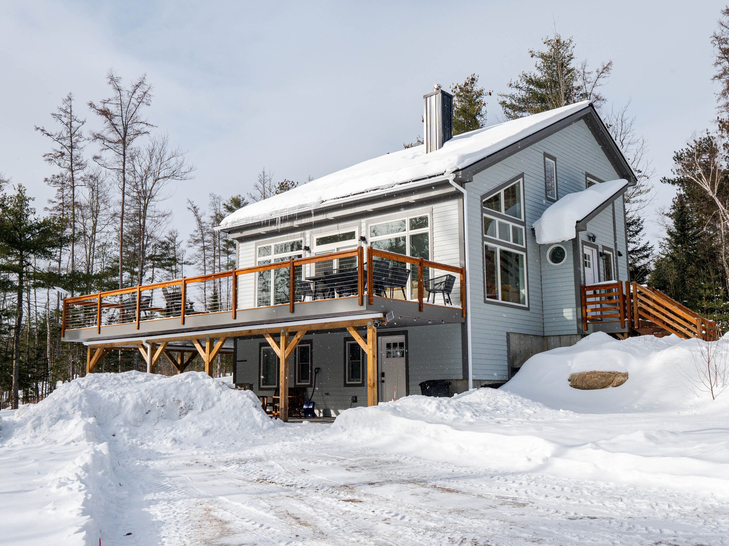 A modern, gray, two-story house with a snow-covered roof and large windows, surrounded by snow, with a wooden deck and stairs, and trees in the background.