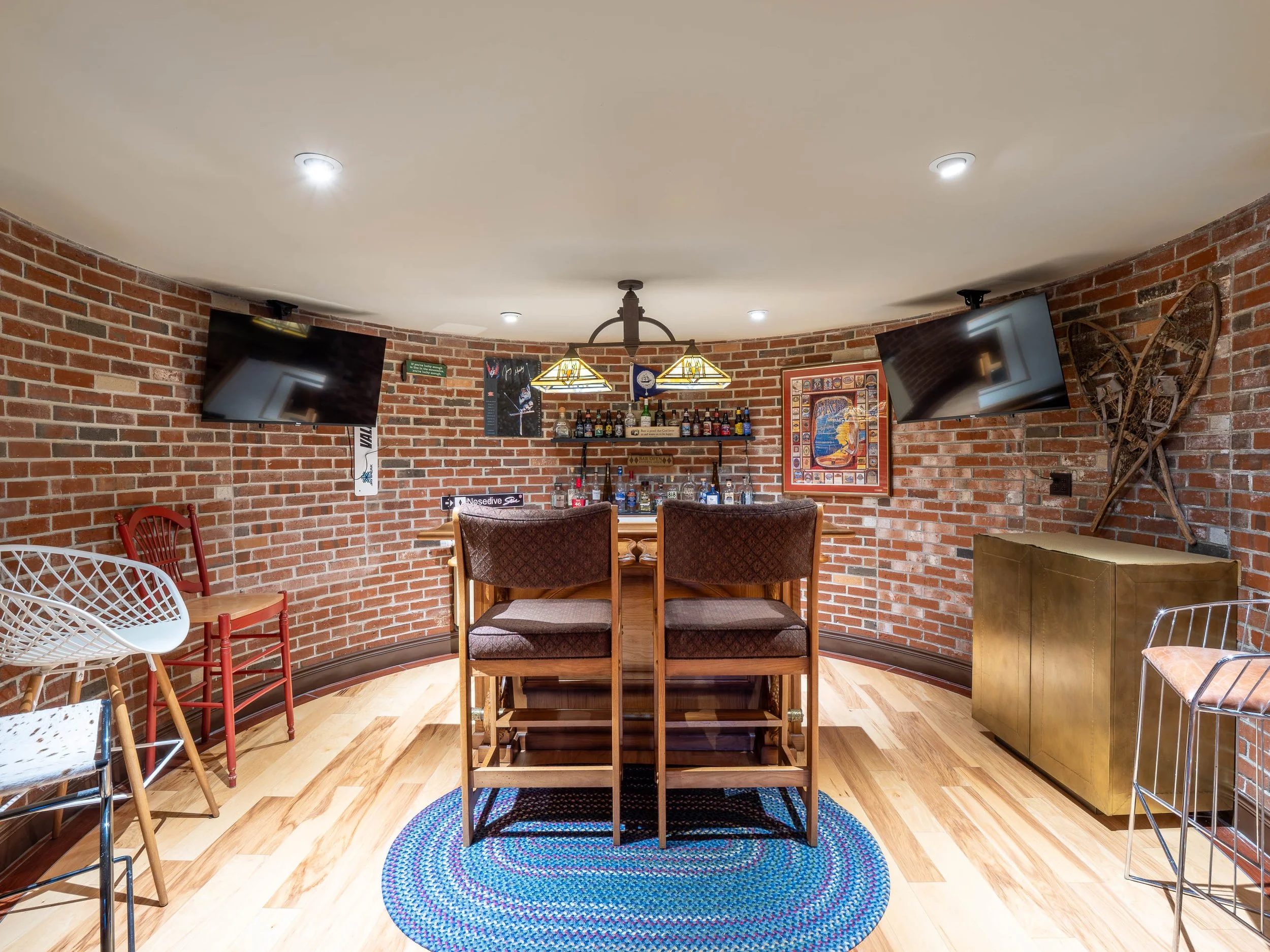 A cozy brick-walled bar with a round wooden table, two upholstered chairs, two bar stools, and a braided rug. Two TV screens are mounted on the walls, and there are decorative items including vintage snowshoes on the right wall, framed posters, artwo