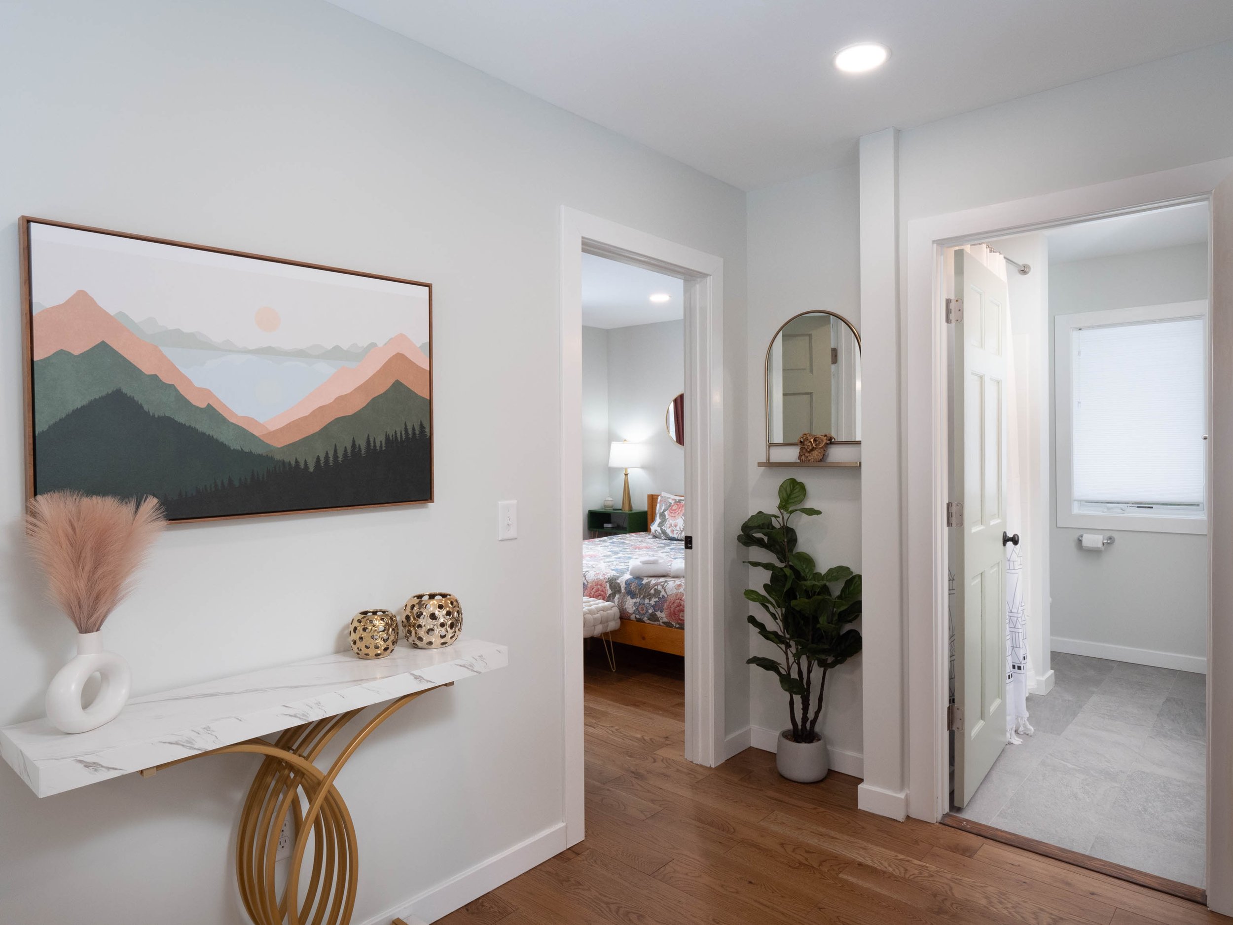 Interior of a modern home hallway with a white console table, framed mountain landscape art, and a potted plant, leading to a bedroom and a bathroom.