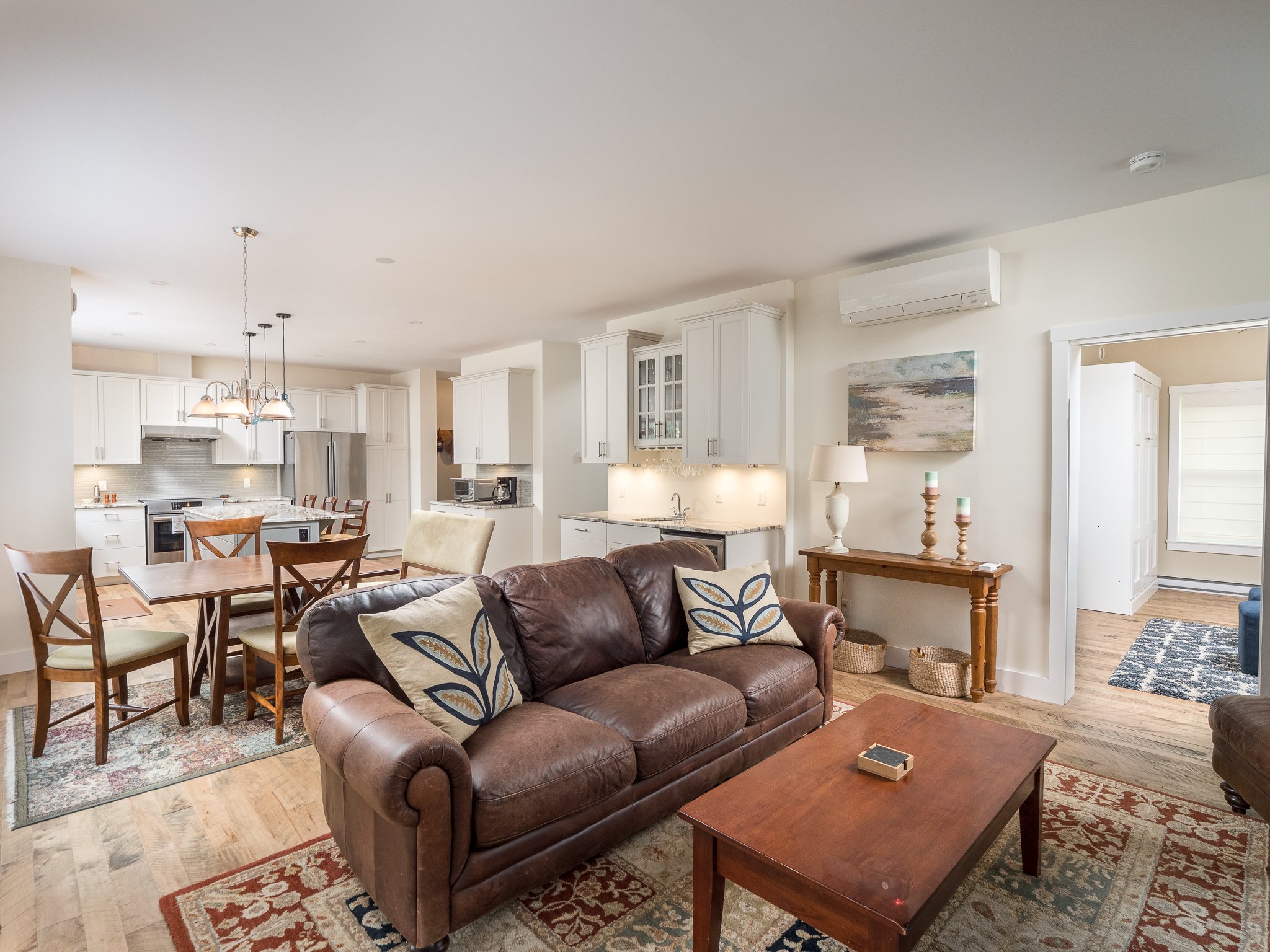 Open-concept living and dining area with a brown leather sofa, wooden coffee table, dining table with chairs, and a modern kitchen. The space features light walls, hardwood floors, and a pendant light fixture.