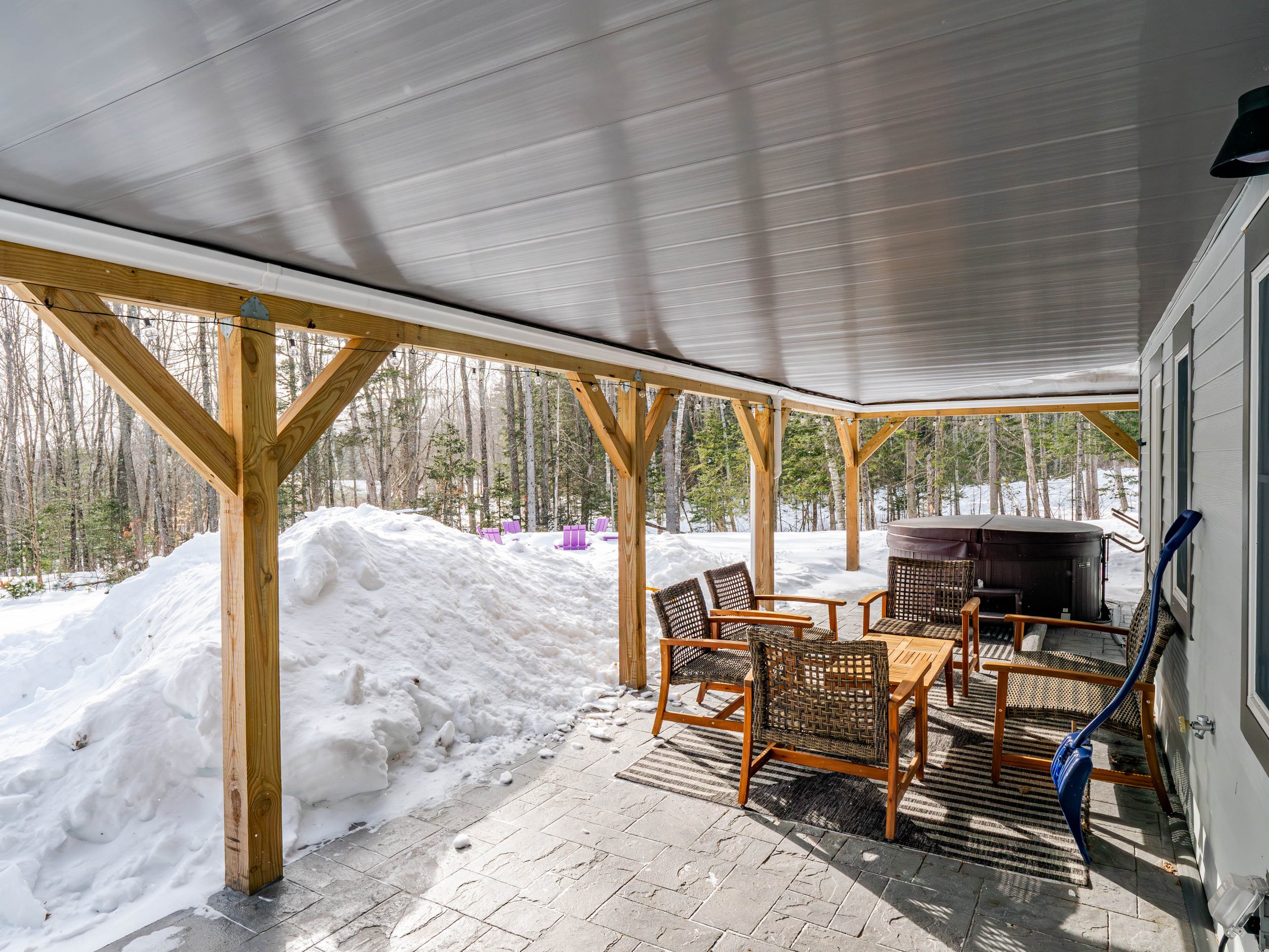 Covered outdoor patio with patio furniture, snow pile, hot tub, and wooded background.