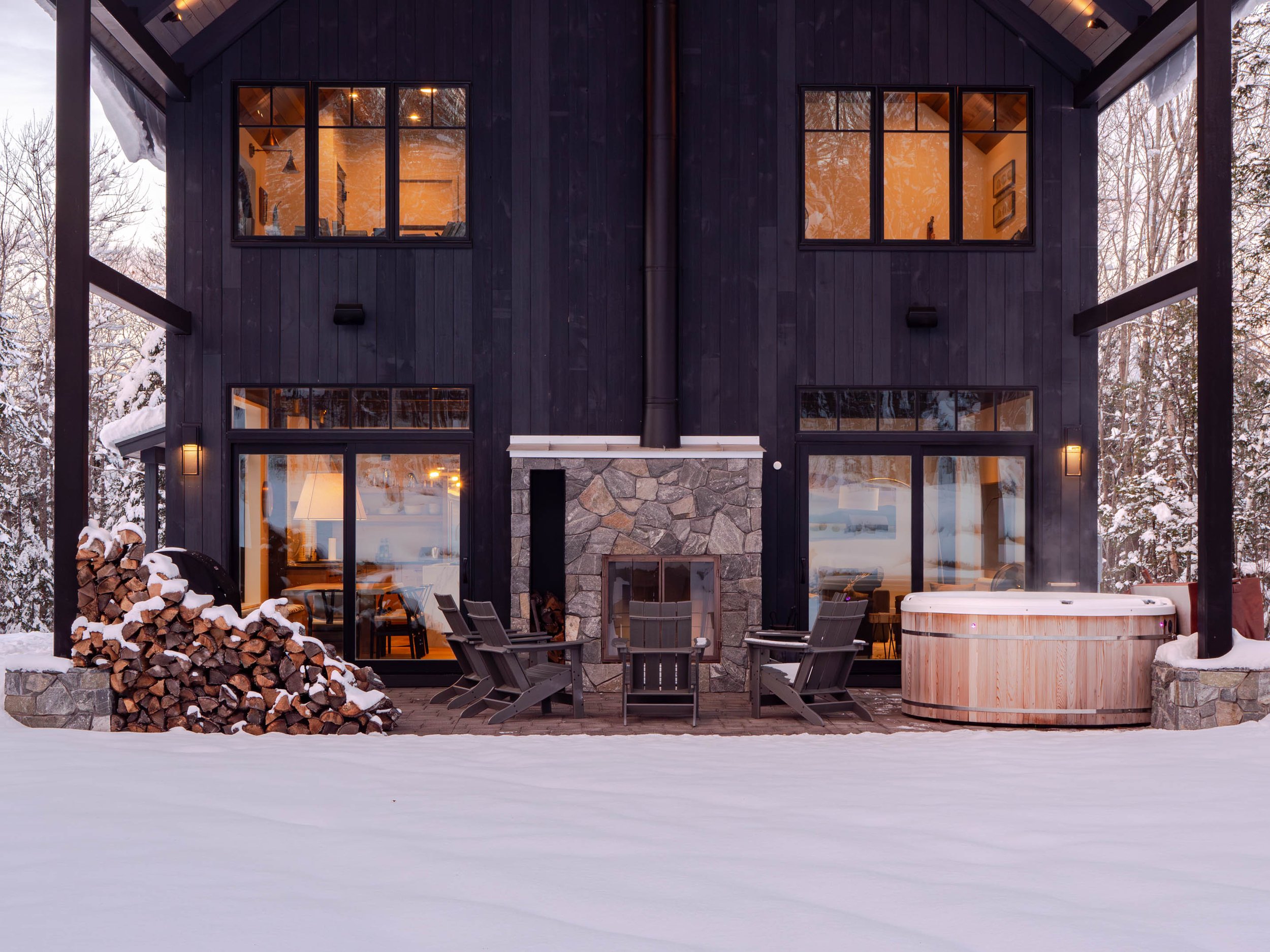 Cozy winter scene of a wooden cabin with large windows, a stacked pile of firewood, outdoor fireplace, seating area, and steaming hot tub, surrounded by snow.