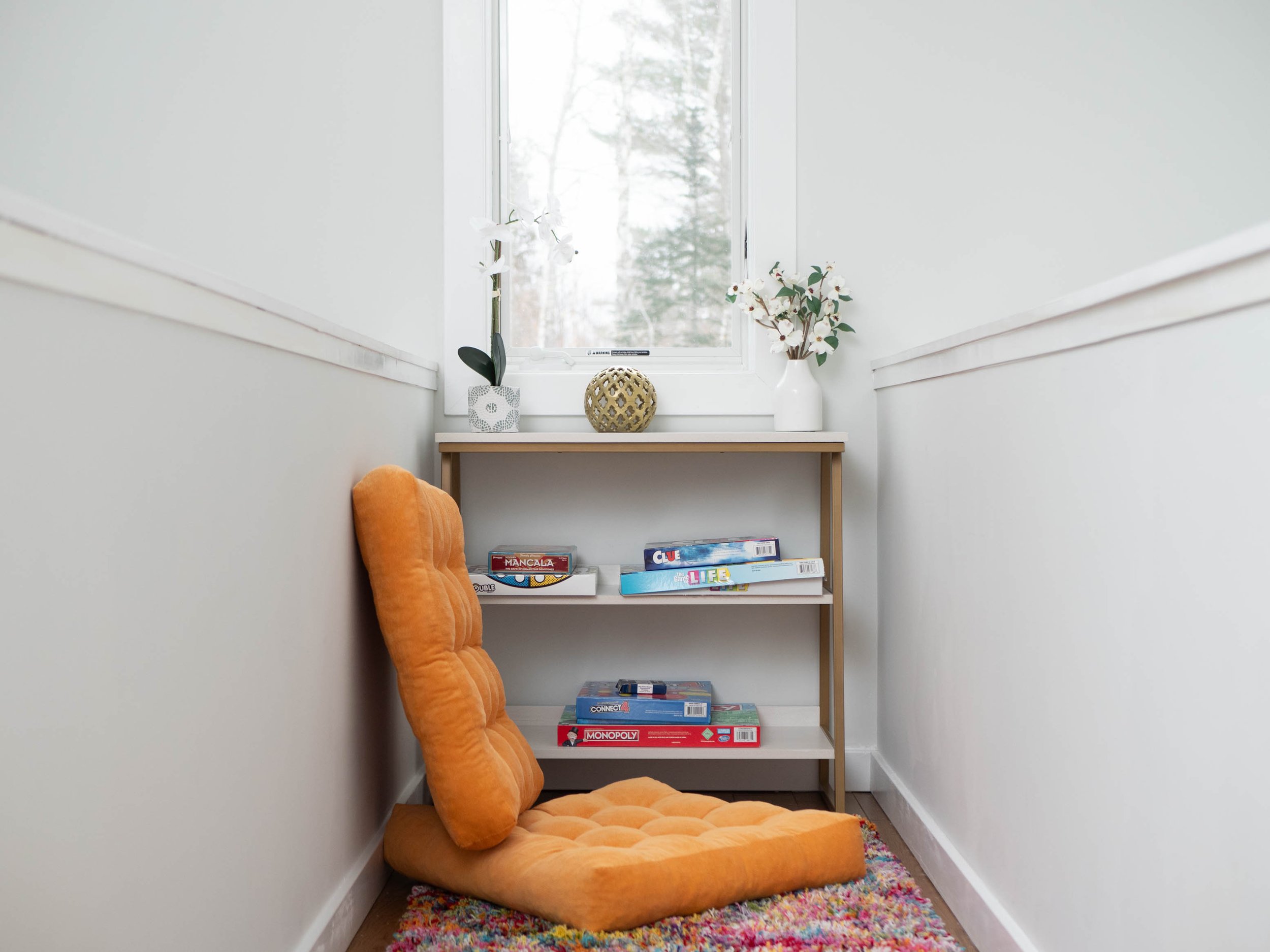 Small cozy nook with white walls, a window, a wooden shelf with board games and decorative flowers, and an orange upholstered floor cushion placed on a multicolored rug.