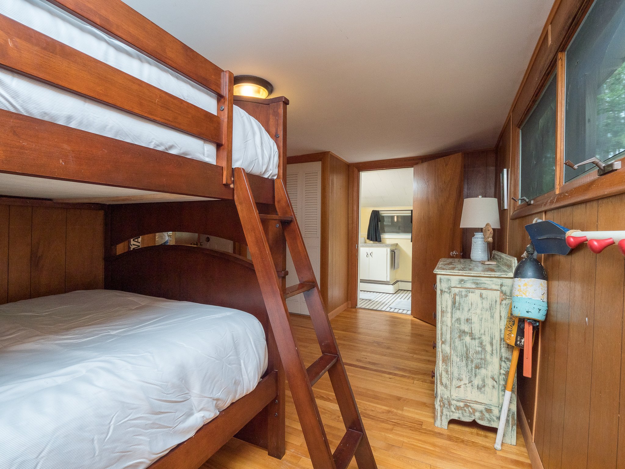 Interior of a wooden cabin room with a bunk bed, rustic dresser, wooden floor, and small lamp; view into a kitchen area.