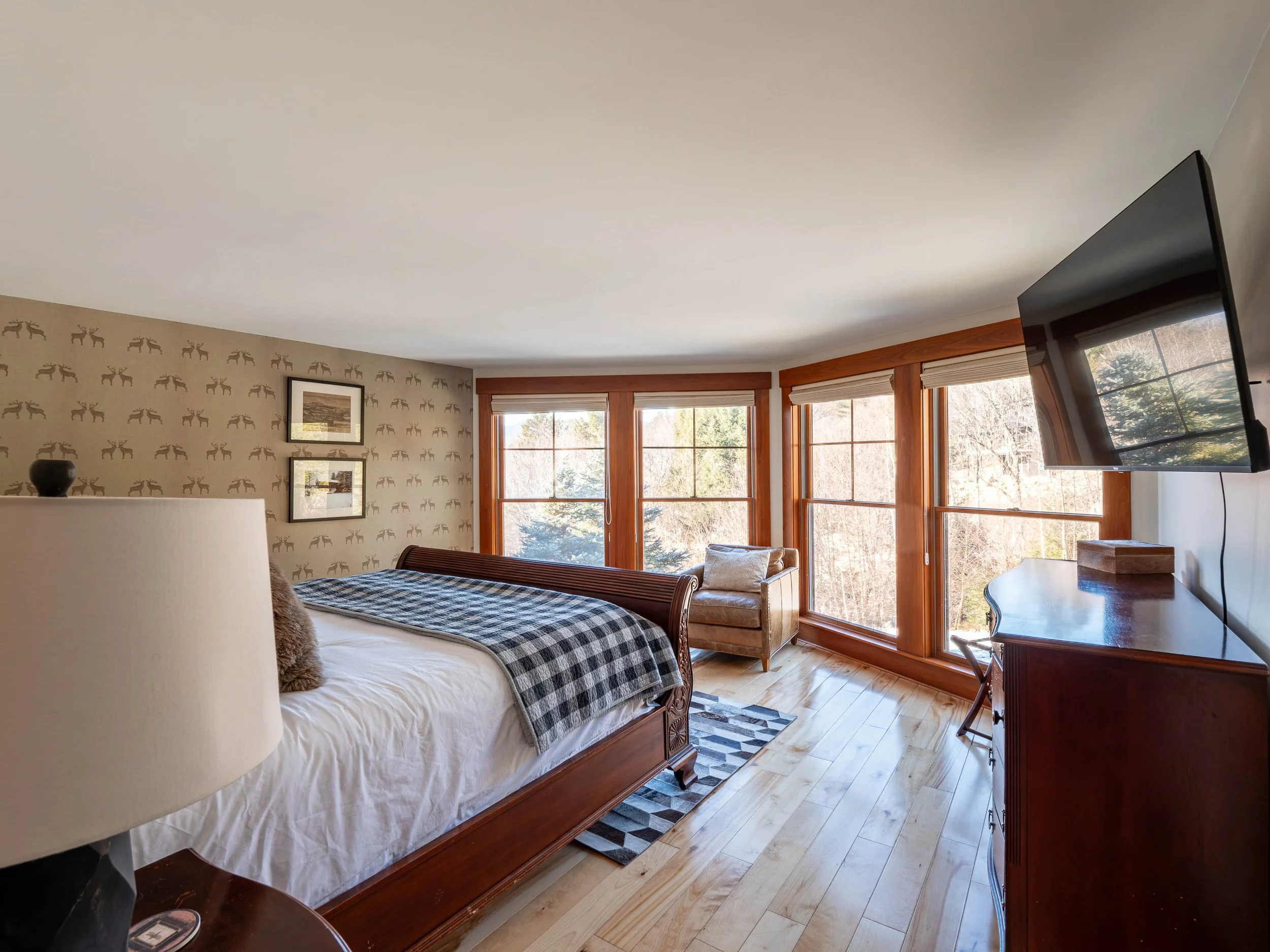 Room with wooden floors, a large bed with white sheets and checkered blanket, a window seat, a wall-mounted TV, and large windows with a view of trees outside.