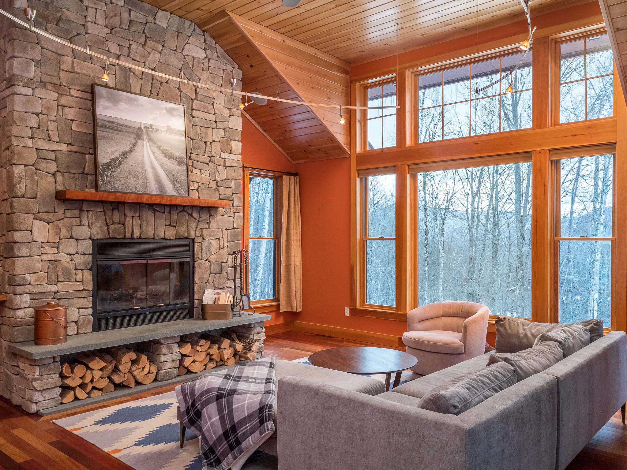 Cozy living room with a stone fireplace, wooden ceiling, large windows, gray sectional sofa, and wooden coffee table.