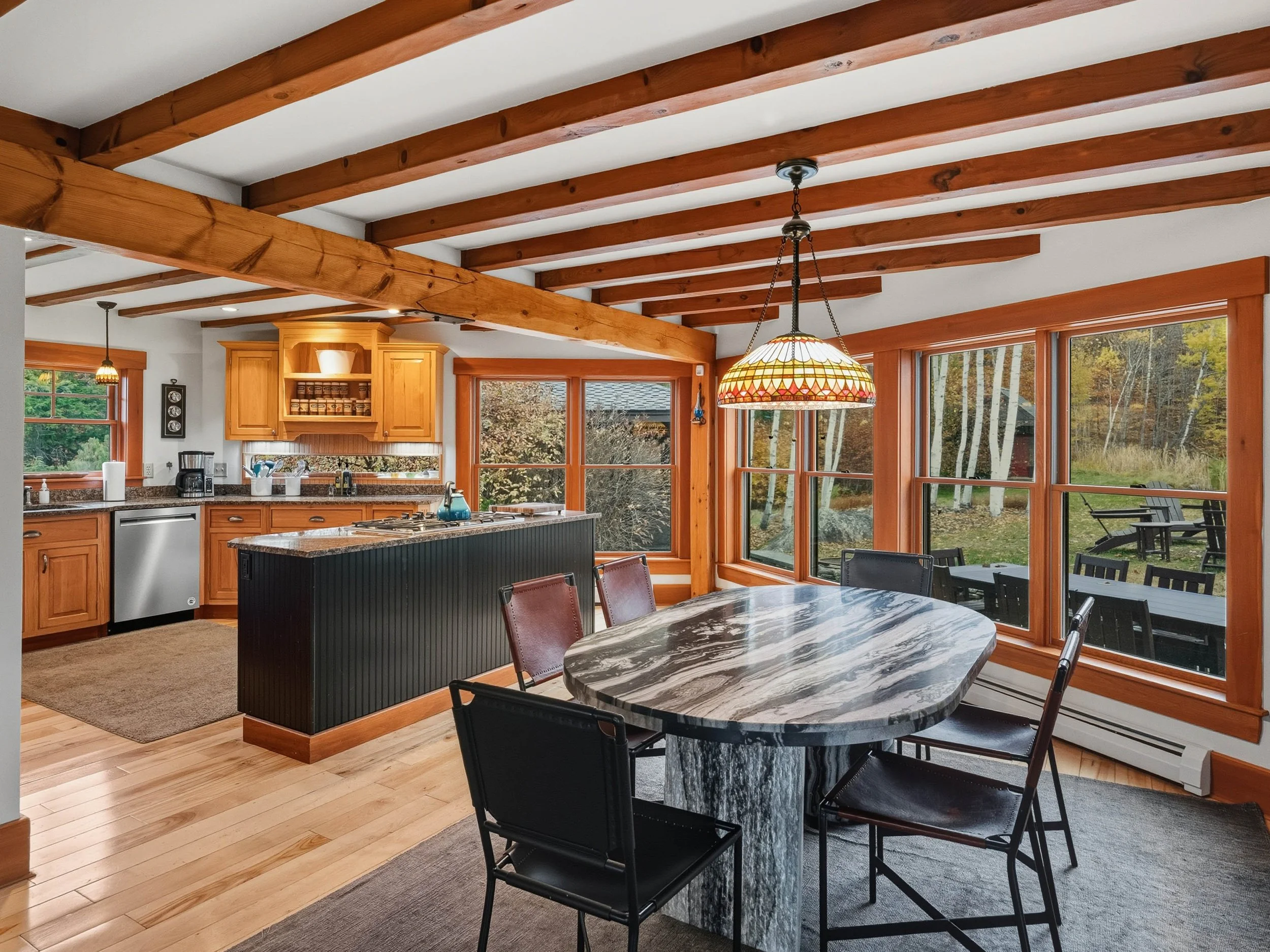 A kitchen and dining area with wooden beams on the ceiling, large windows showing an outdoor patio and trees, a marble dining table with black chairs, a black kitchen island, and wooden kitchen cabinets.