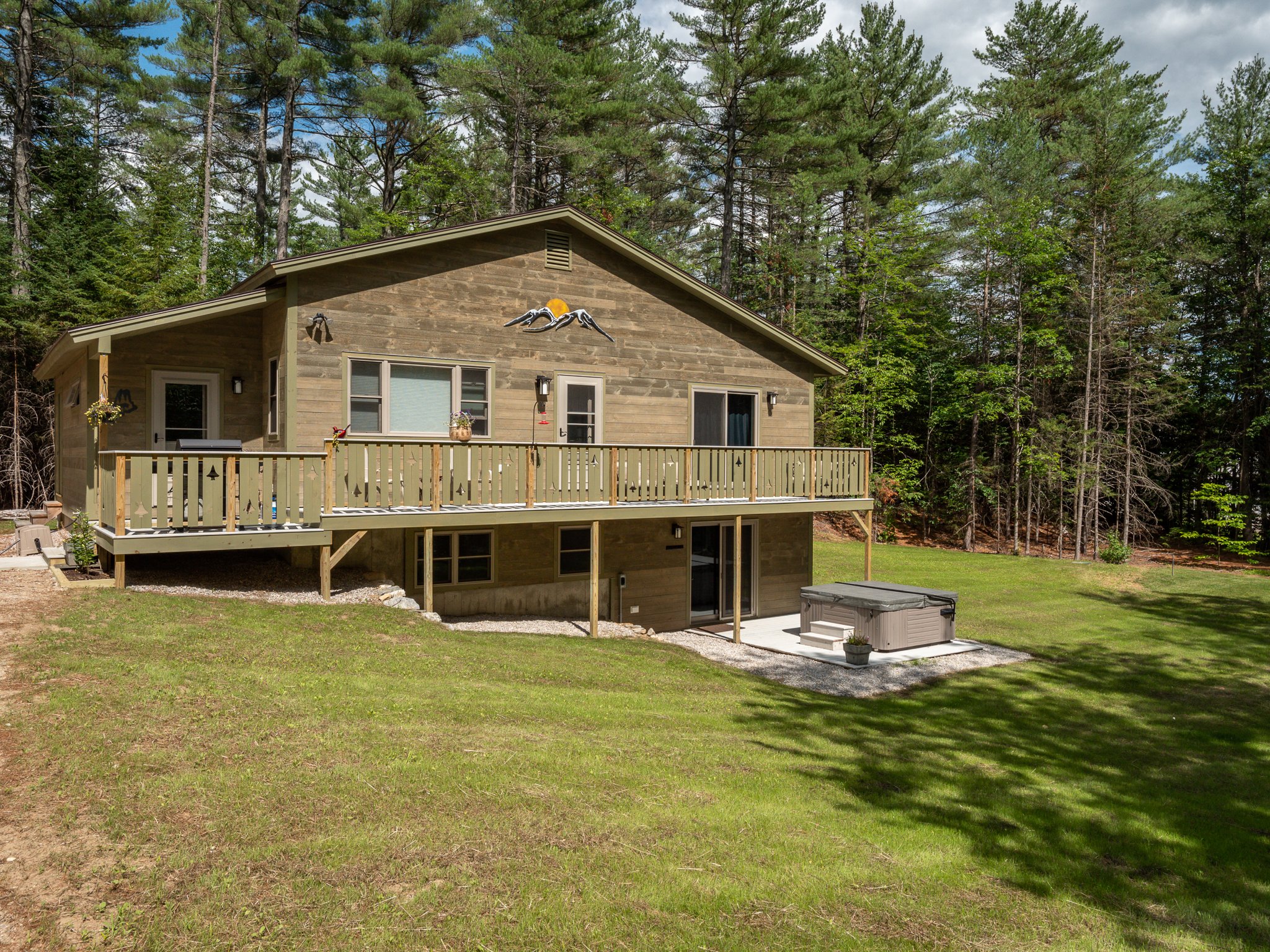 Rural two-story house with wooden deck, surrounded by pine trees, featuring an outdoor hot tub on a concrete patio and a grassy lawn.