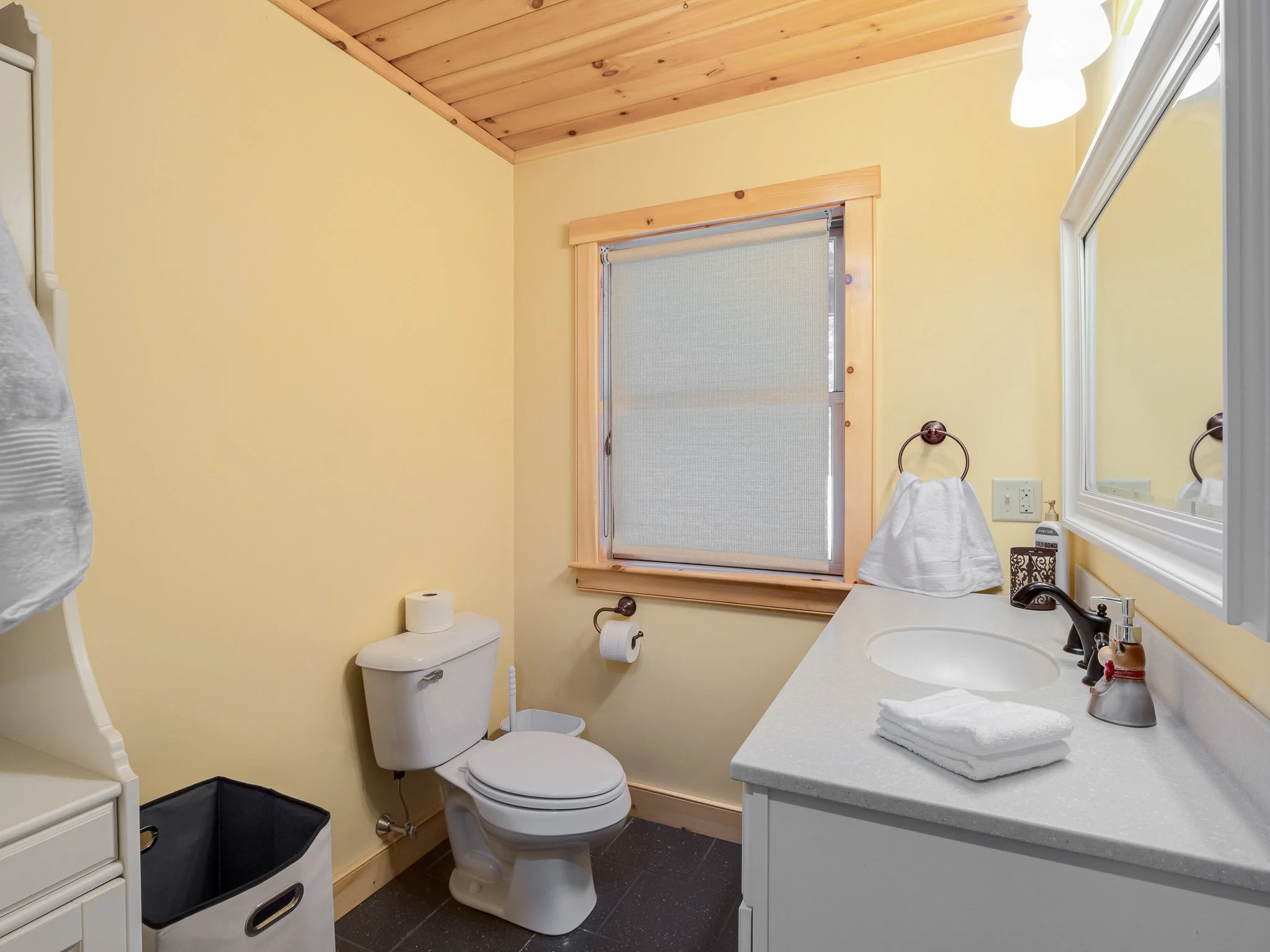 Bathroom with yellow walls, white toilet, white countertop with sink, folded towels, soap dispenser, window with rolled-up shade, and black tile floor.