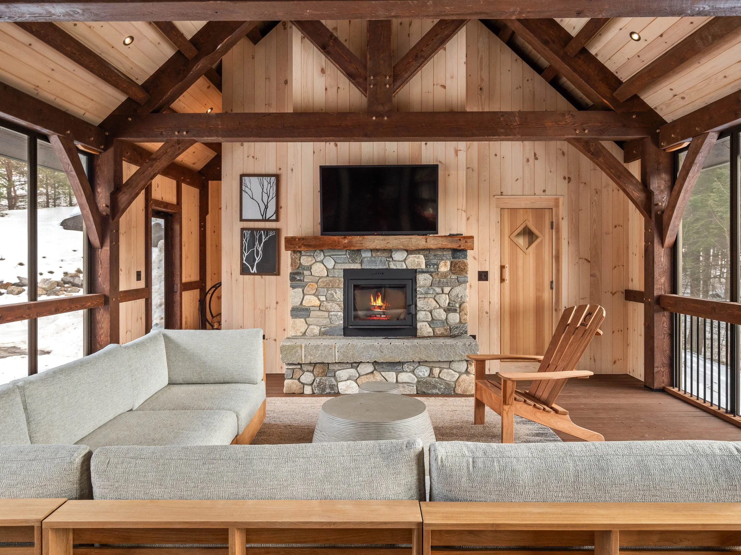 Cozy living room with wood-paneled walls and ceiling, a stone fireplace with a flat-screen TV above, a beige sectional sofa, a wooden armchair, and a round coffee table. Large windows show a snowy landscape outside.