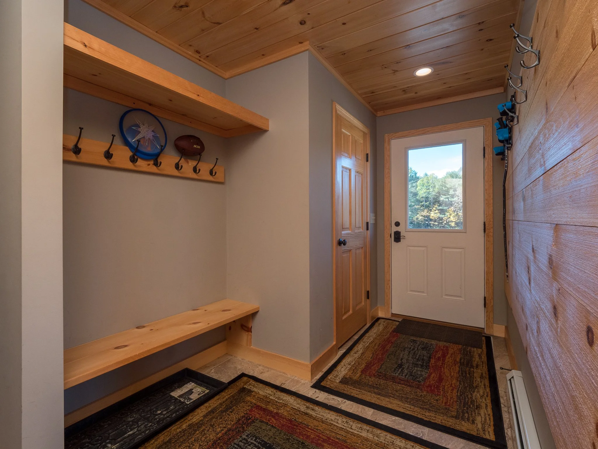 A rustic mudroom with wooden walls and ceiling, featuring a bench and hooks for storage. The space includes a door with a glass window, two rugs on the floor, and sports equipment like a basketball and frisbee on a shelf.