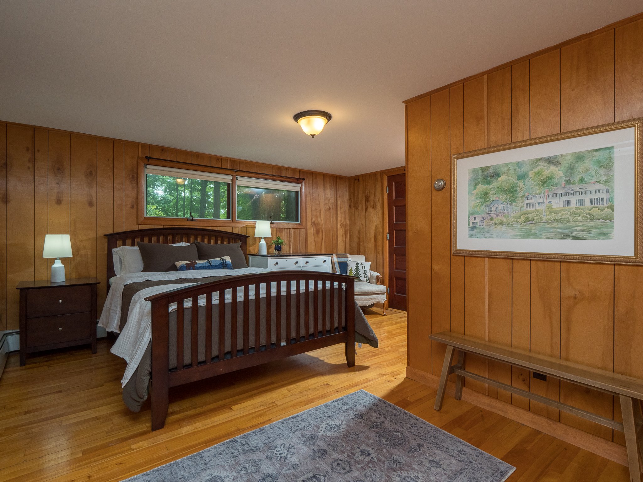 Bedroom with wood paneling, a wooden bed frame, bedside tables with lamps, a dresser, artwork on the wall, and a bench.