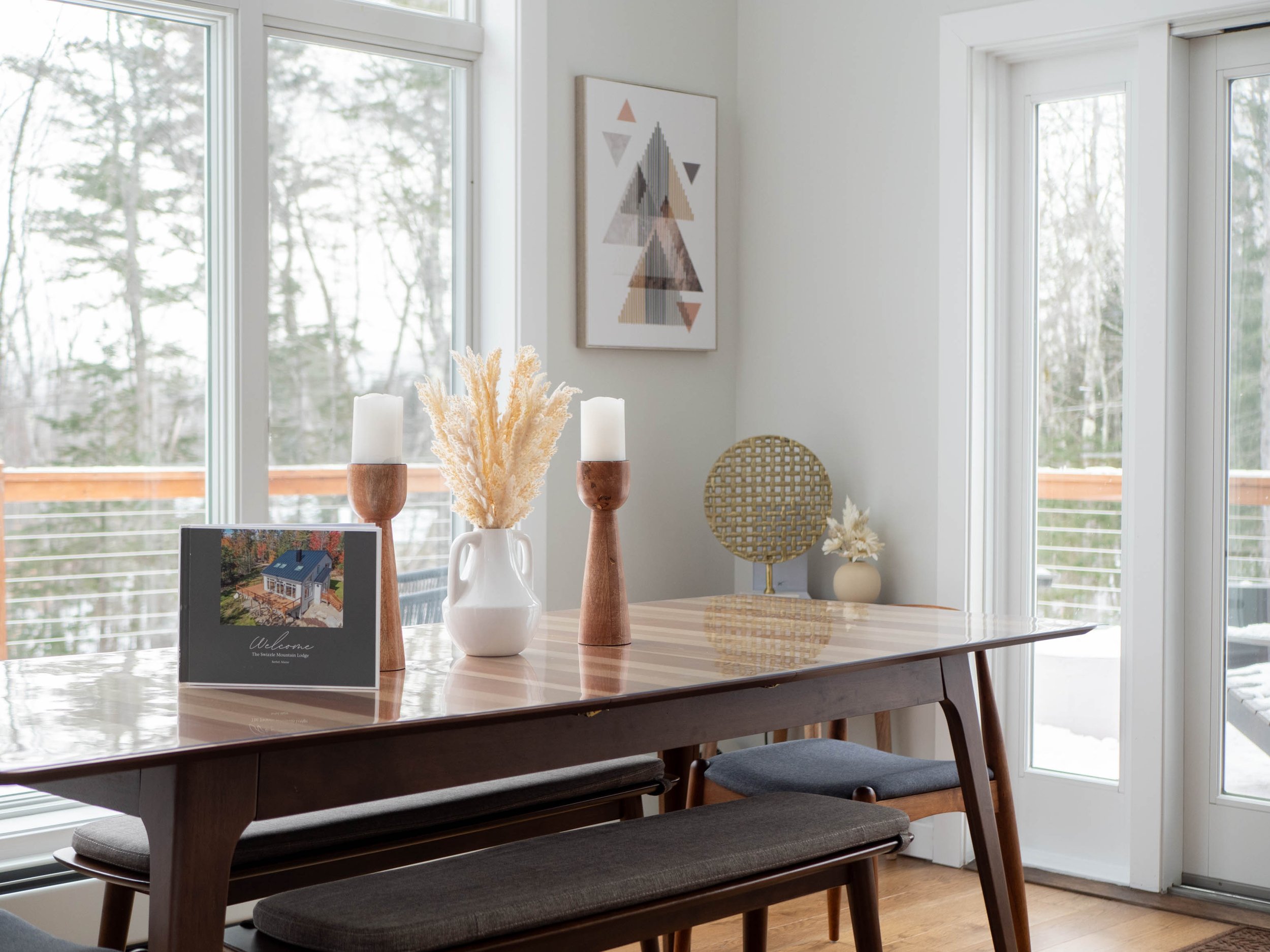 Decorated dining table with candles, a white vase with dried flowers, a framed photograph, and a decorative gold round piece, in a room with large windows showing a snowy outdoor scene.
