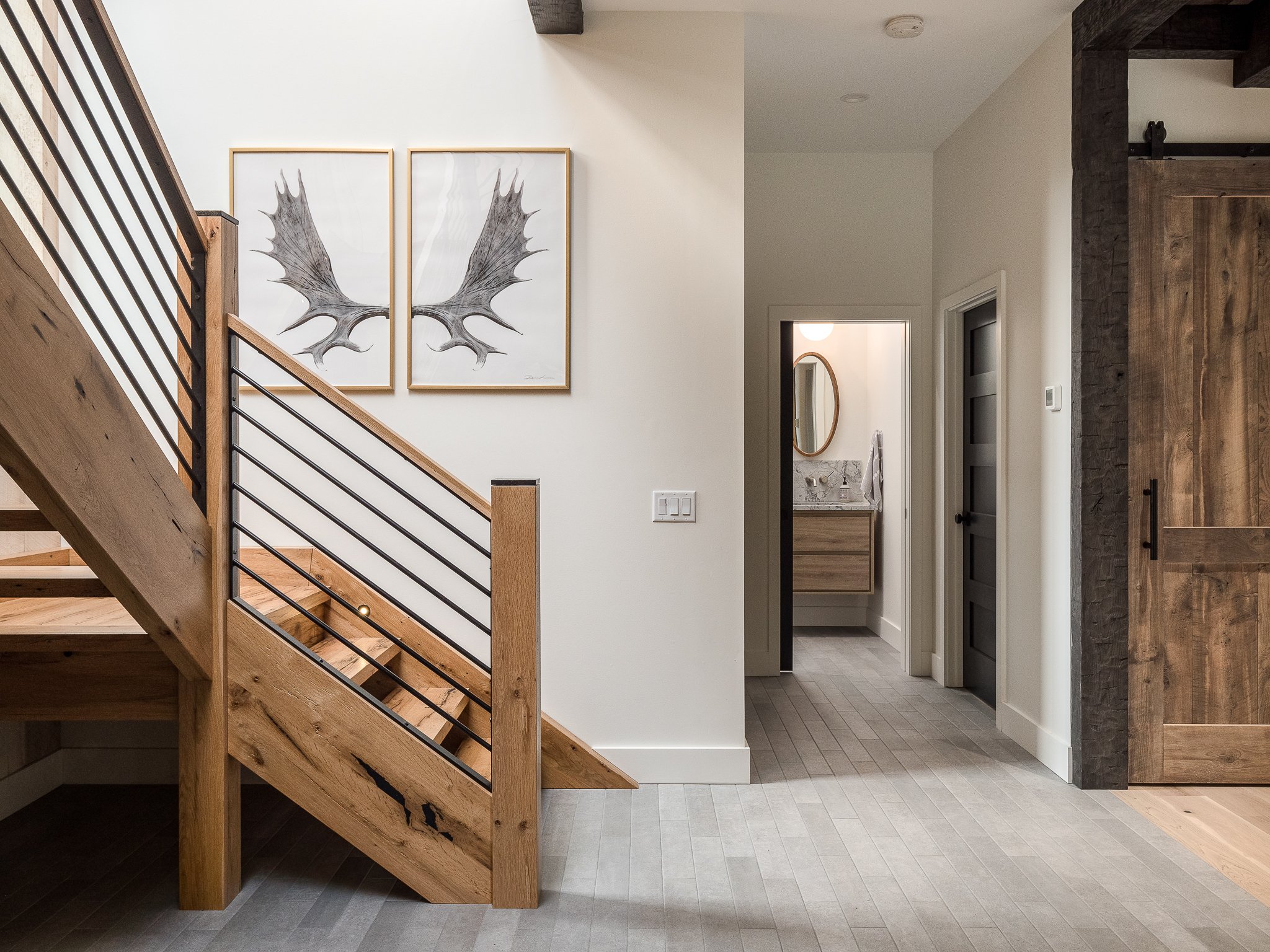 Modern hallway with wood staircase, antler artwork, sliding barn door, and view into bathroom.