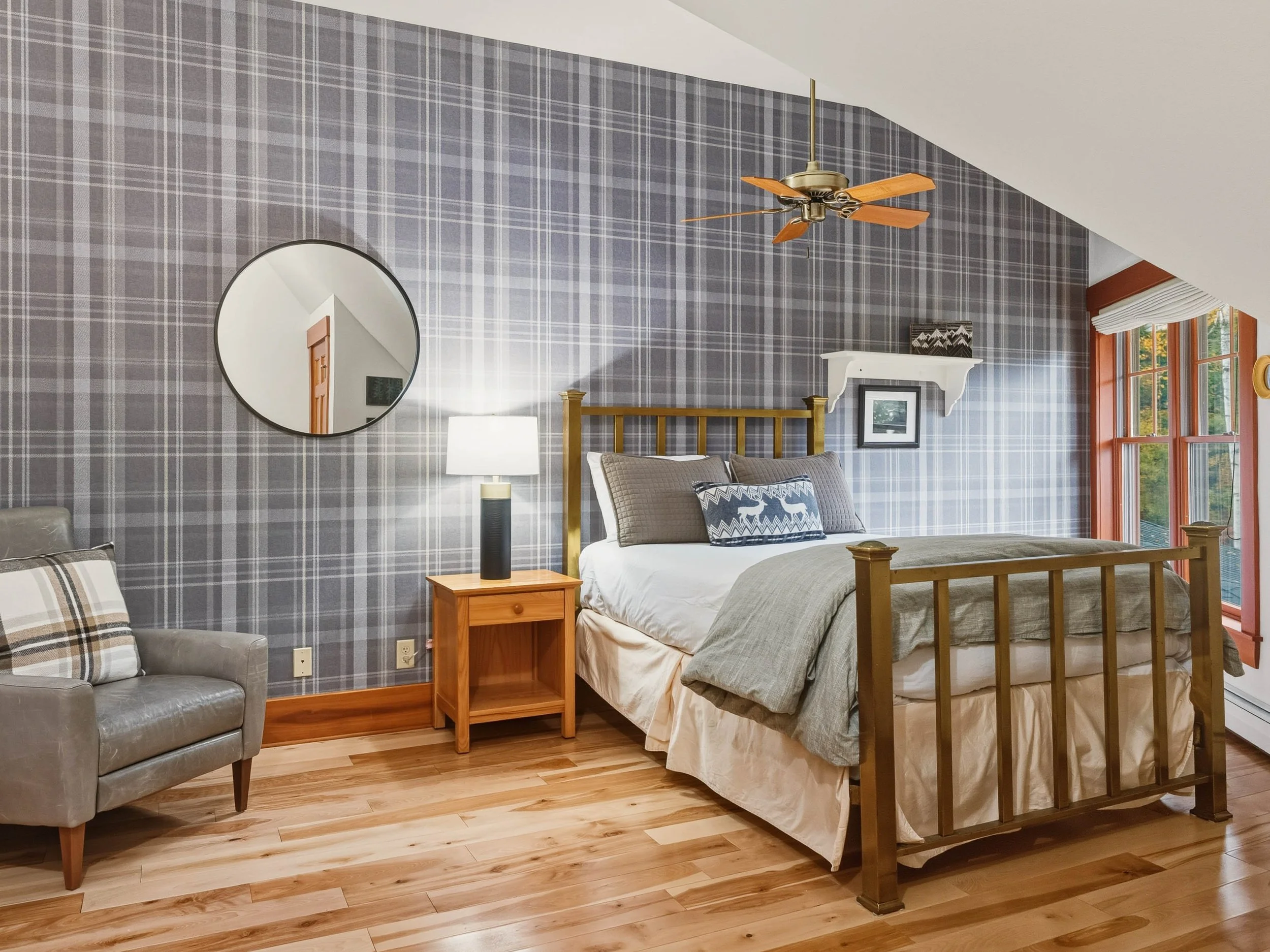 A bedroom with a patterned gray and blue plaid accent wall, a wooden bed with gray bedding, a gray armchair with a plaid pillow, a wooden nightstand with a black and white lamp, and a ceiling fan with wooden blades.