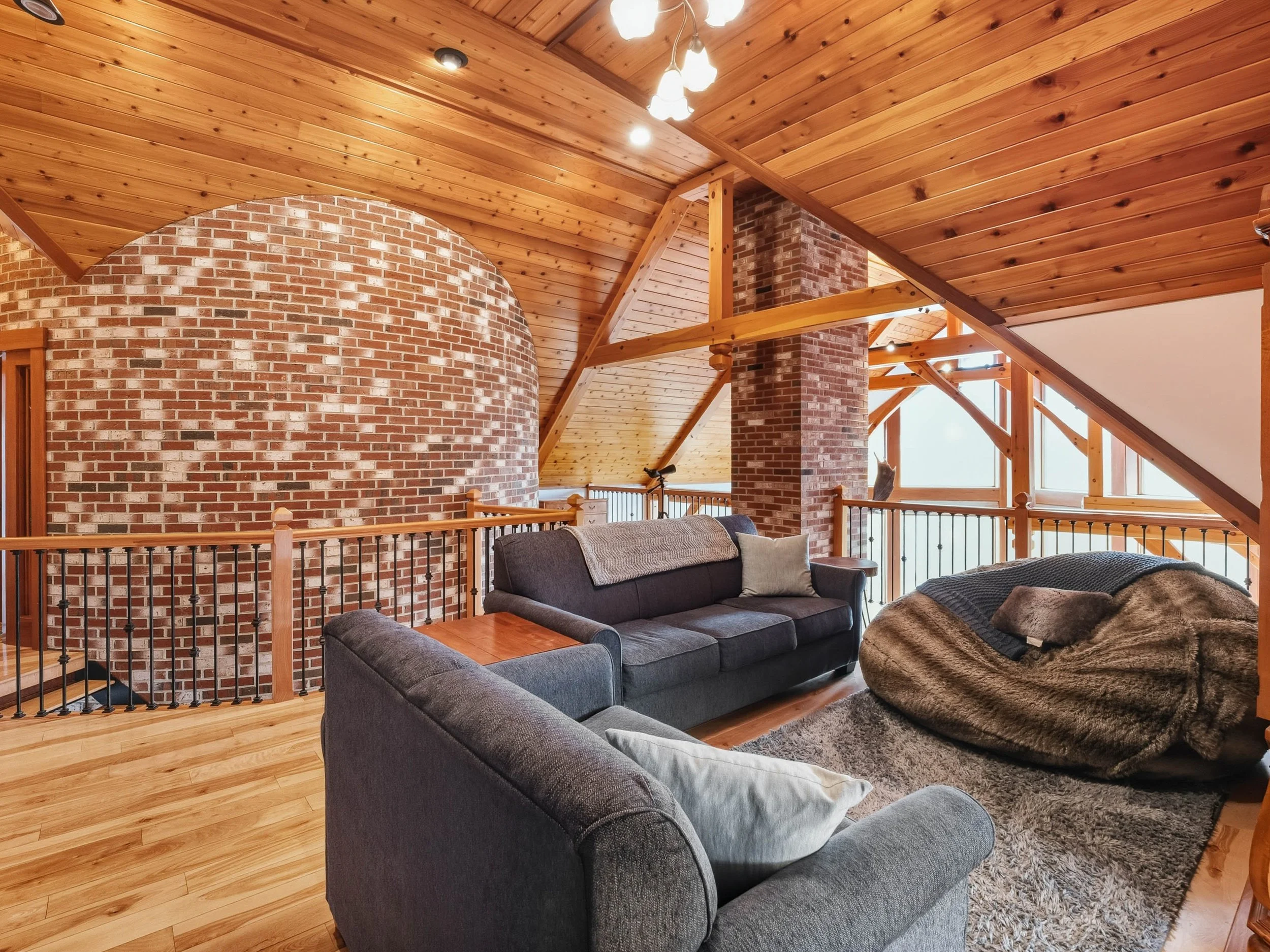 Living room with wood-paneled sloped ceiling, brick wall, large windows, and sofas with blankets and pillows.