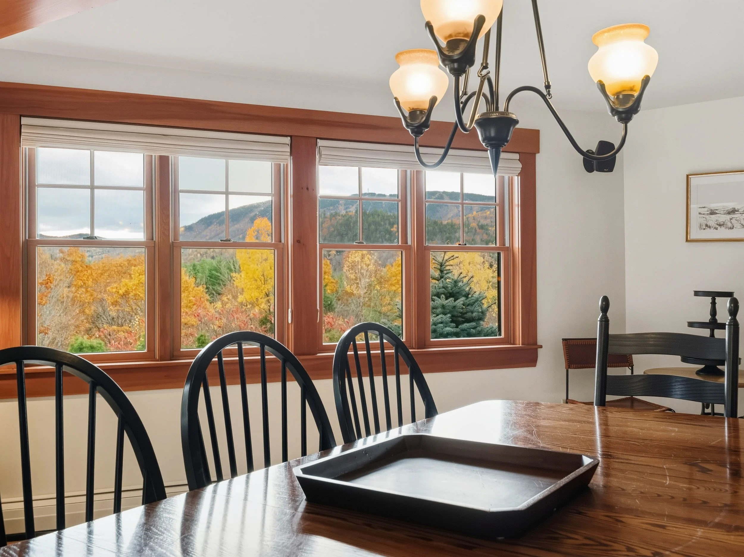 Dining room with large window view of autumn trees and mountains, wooden table with black chairs, chandelier lighting.