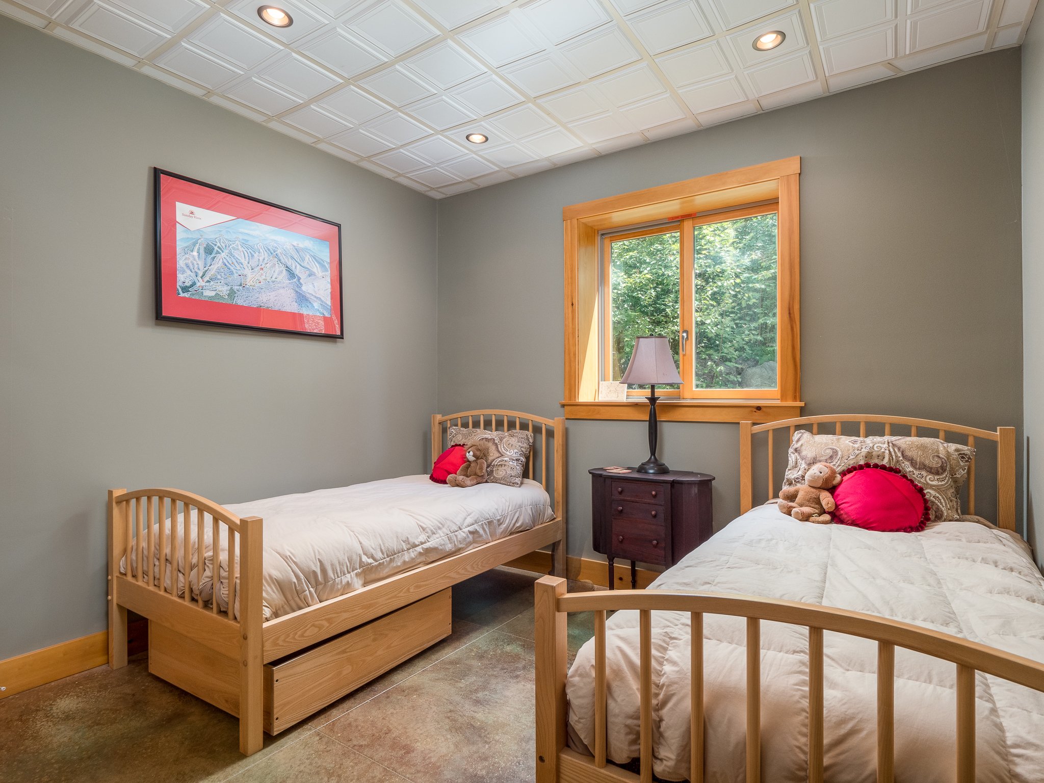 Cozy bedroom with two twin beds, light wood frames, neutral bedspreads, a nightstand with a lamp between them, a bear and pillow on one bed, a window with green views, and a framed artwork on a gray wall.