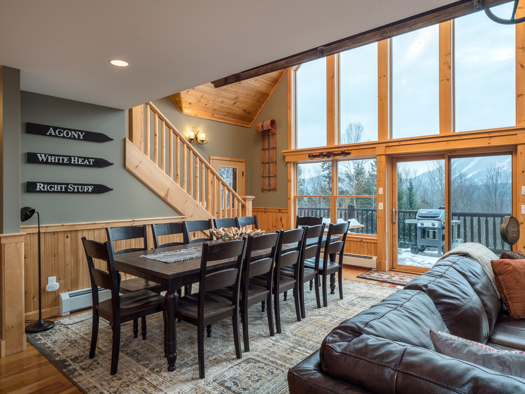 Cozy dining room with a long wooden table, chairs, and a leather couch. Large glass windows offer a view of a balcony and snowy landscape. Wooden interior accents and a staircase are visible.