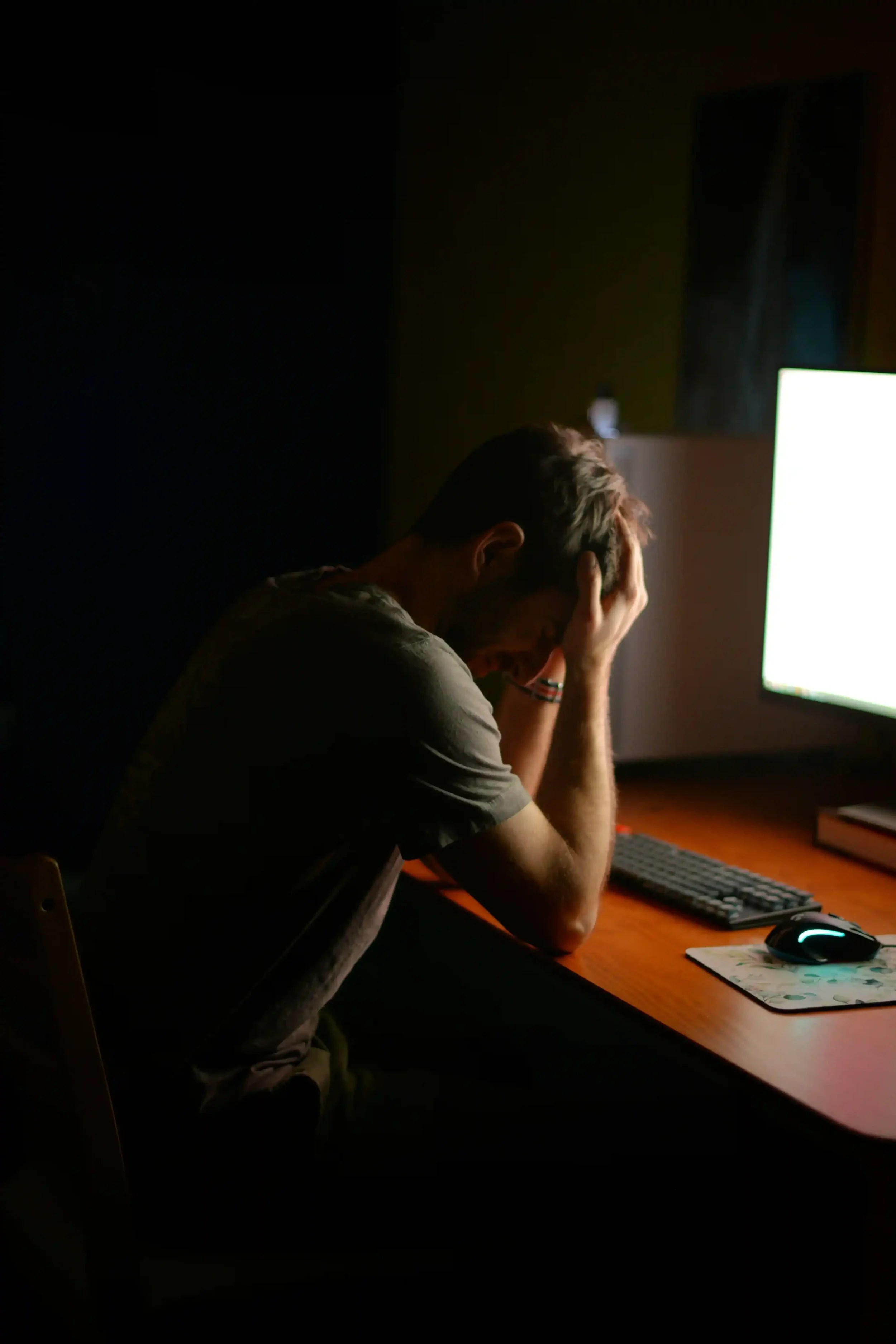 A man sits at a desk in a dimly lit room, his head resting in his hands in a gesture of exhaustion or stress.