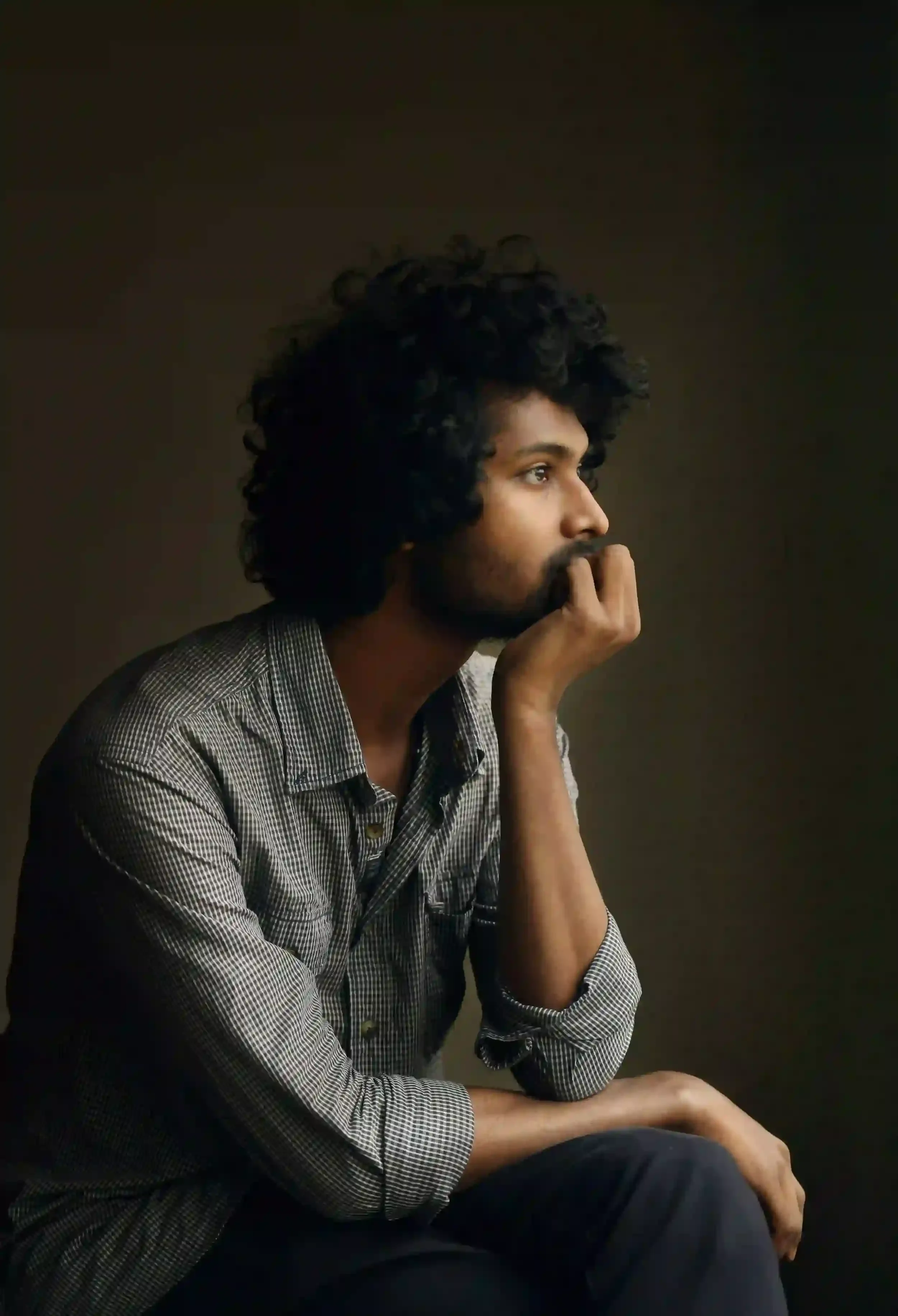 A side profile portrait of a young man with dark, curly hair and a beard, sitting in a thoughtful or pensive pose against a dark, moody background
