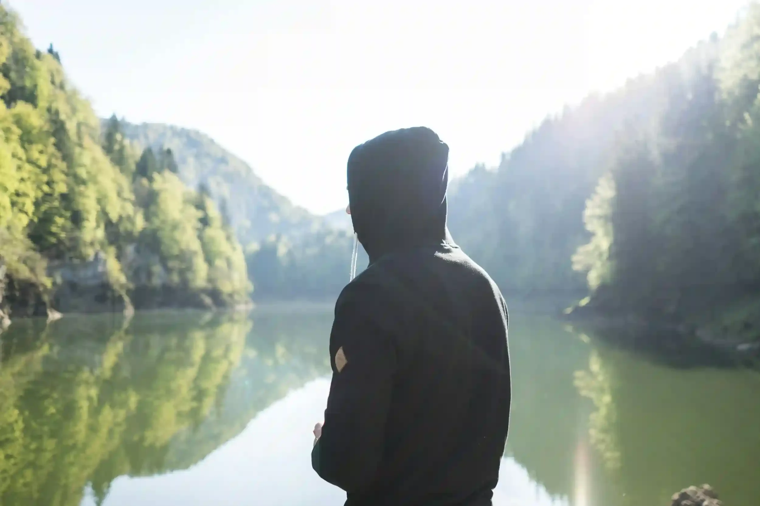 man with back turned wearing a black hooded jacket overlooking a lake with a mountainous background