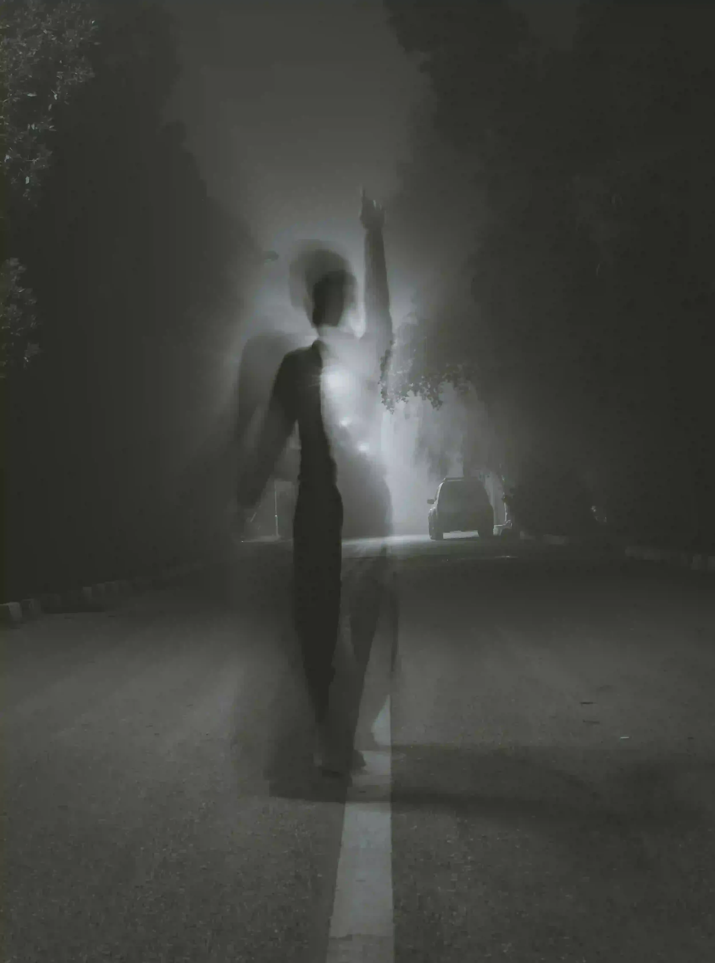 A black and white, long-exposure photograph of a person standing in the middle of a foggy road at night.