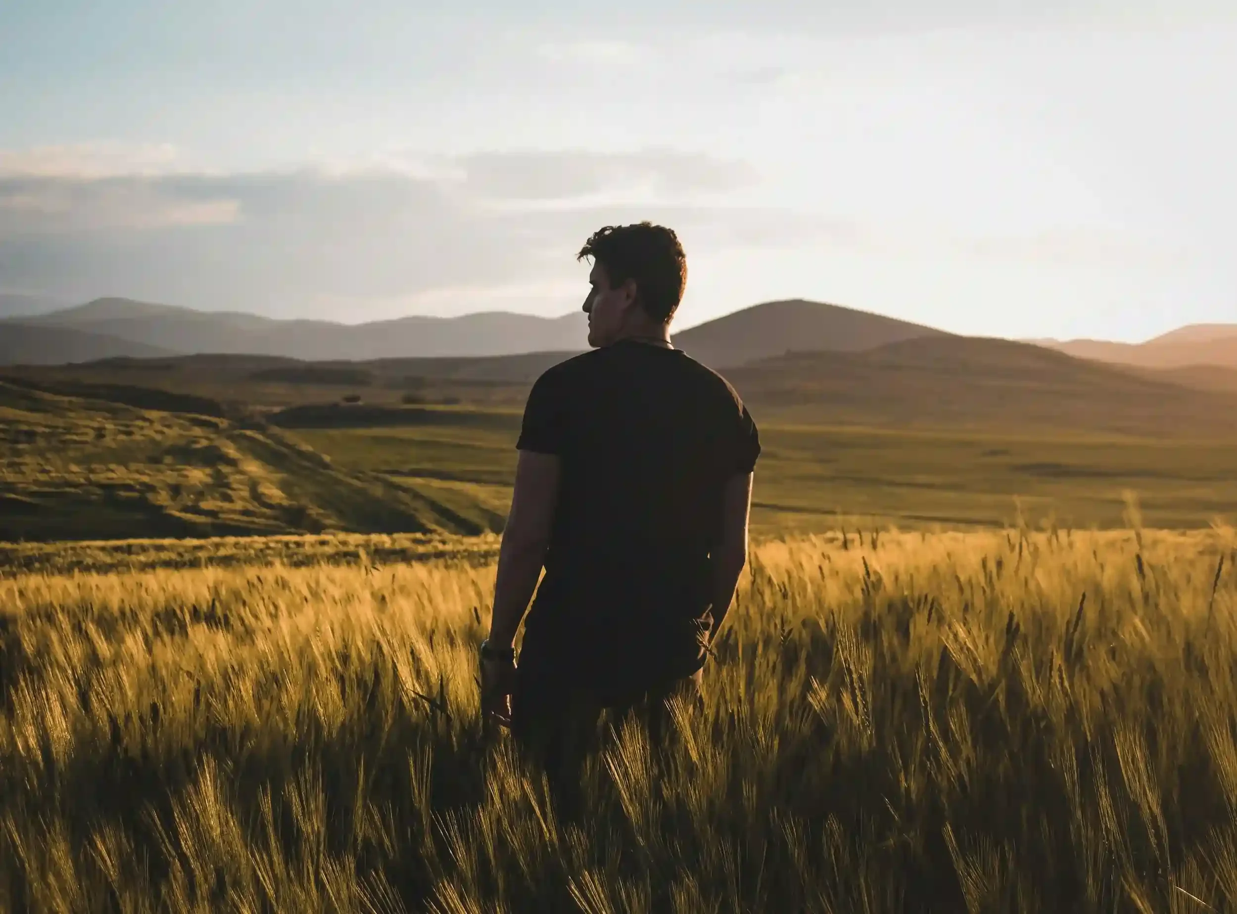man with back turned and side profile showing standing in field of tall grass with mountain view in the distance
