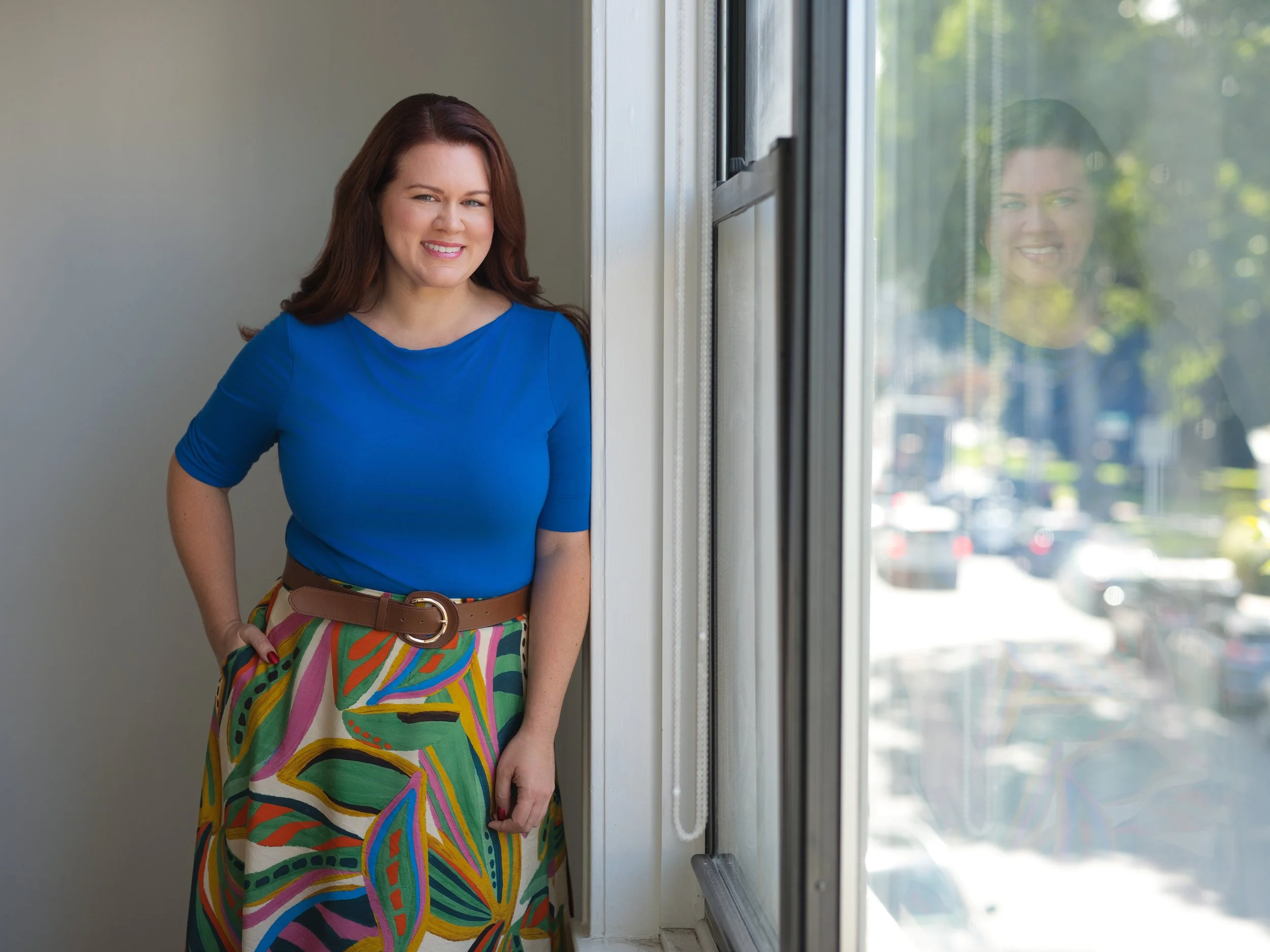A woman with long, wavy auburn hair wearing a bright blue top and a colorful, patterned skirt, standing indoors near a window with greenery visible outside, smiling at the camera.
