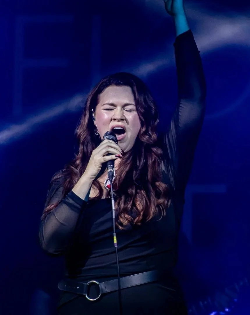 A woman singing passionately on stage with one arm raised and eyes closed, holding a microphone in her other hand, illuminated by stage lights.