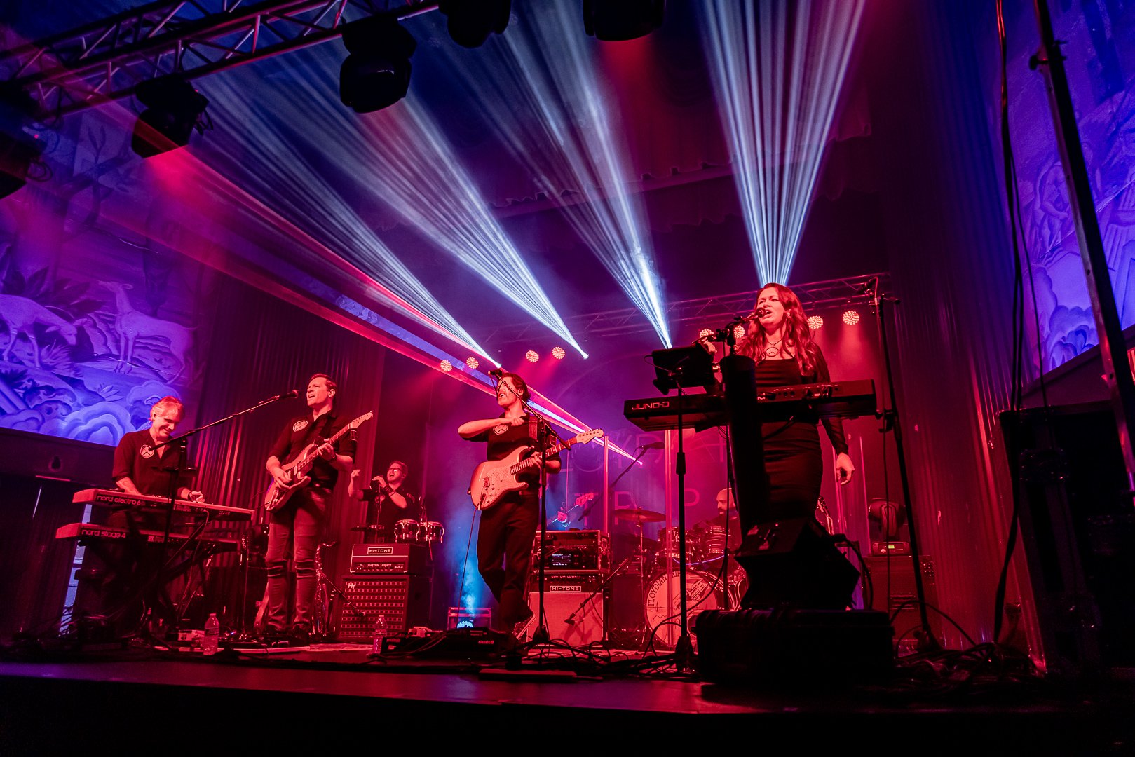 A band of five musicians performing on stage with colorful lighting and a painted backdrop featuring nature scenes.