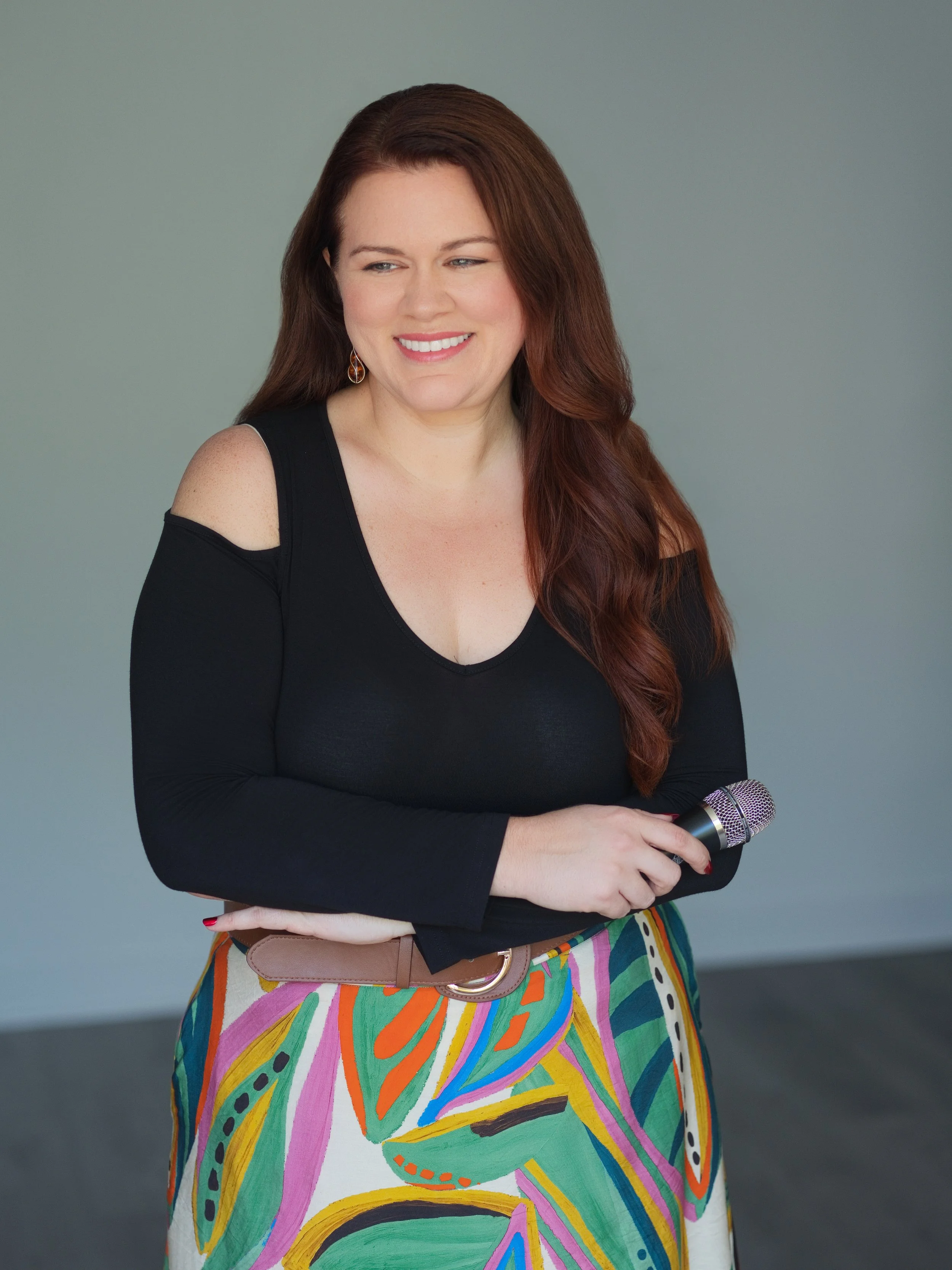 A woman with long, wavy auburn hair smiling, holding a microphone, wearing a black top with cut-out shoulders and a colorful abstract-patterned skirt, standing against a plain gray background.