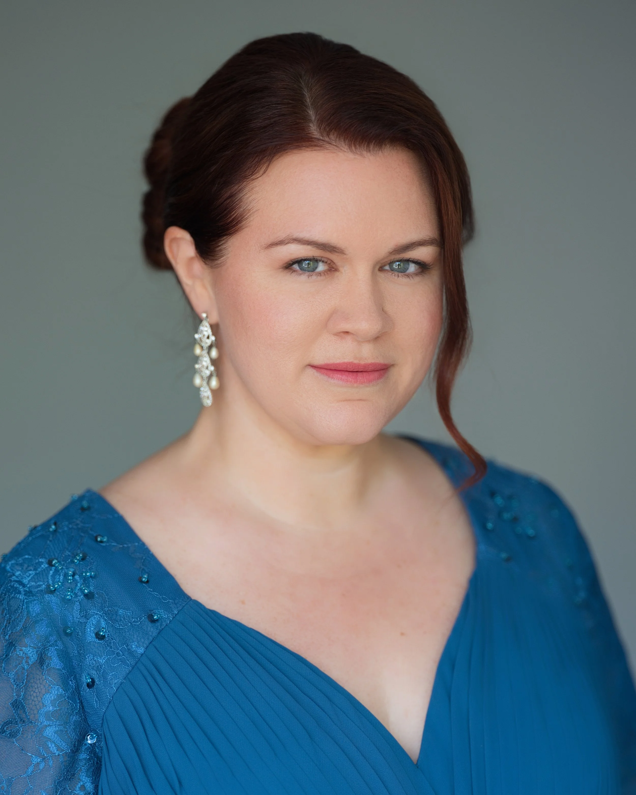 A woman with auburn hair, in an updo, wearing dangling pearl earrings, and a blue dress with lace and bead embellishments, facing slightly to the right, against a plain gray background.