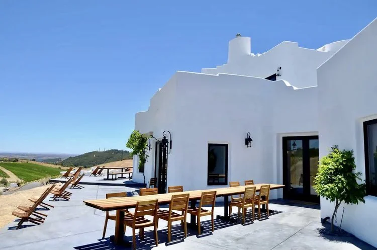 White stucco house with outdoor patio including wooden chairs and table, overlooking a scenic landscape under a clear blue sky.