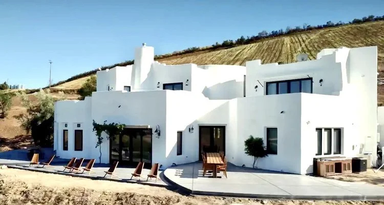 Modern white house with flat roofs and large windows, surrounded by a vineyard on a hillside, featuring outdoor seating and a patio.