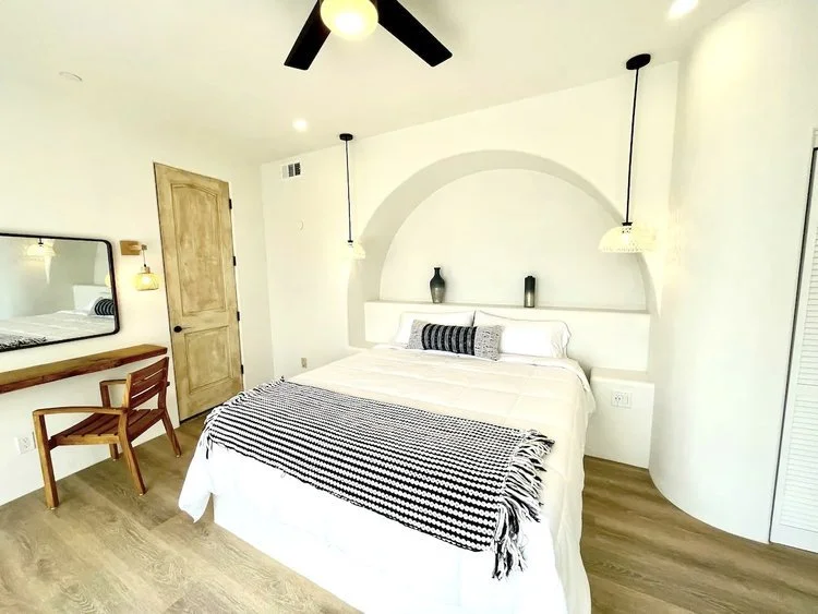 Minimalist bedroom with white walls, wood flooring, a queen-sized bed, black and white patterned throw, wooden door, and a wooden chair next to a wall-mounted mirror. Two pendant lights hang over bedside shelves.