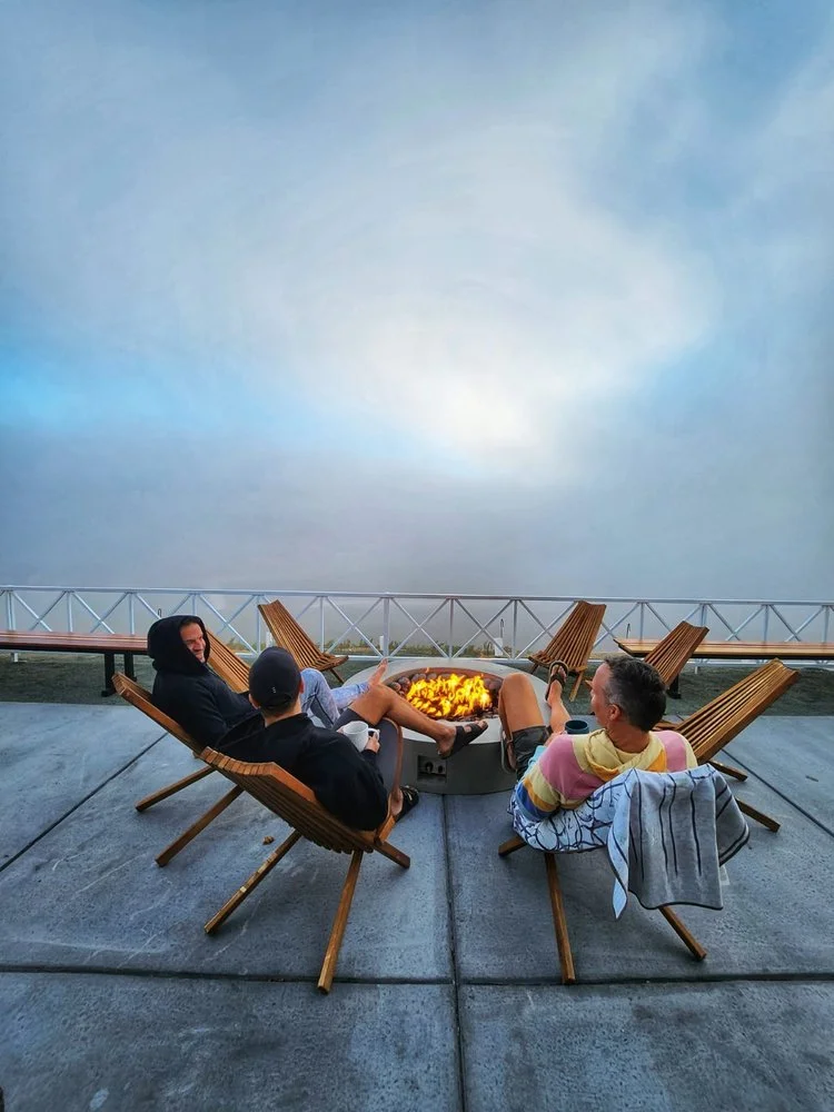 Three people sitting on wooden chairs around a fire pit on a concrete patio, with a cloudy sky and railing in the background.