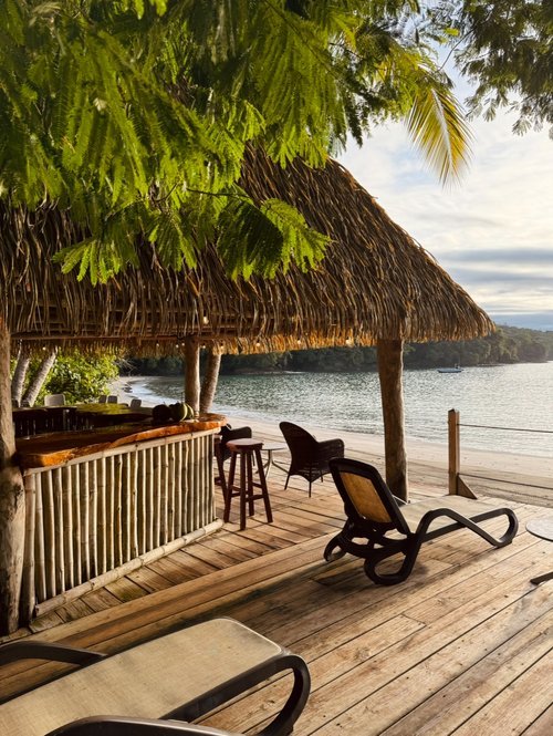 Tropical beachside bar with straw roof, wooden deck, sun loungers, and stools, overlooking a calm ocean.