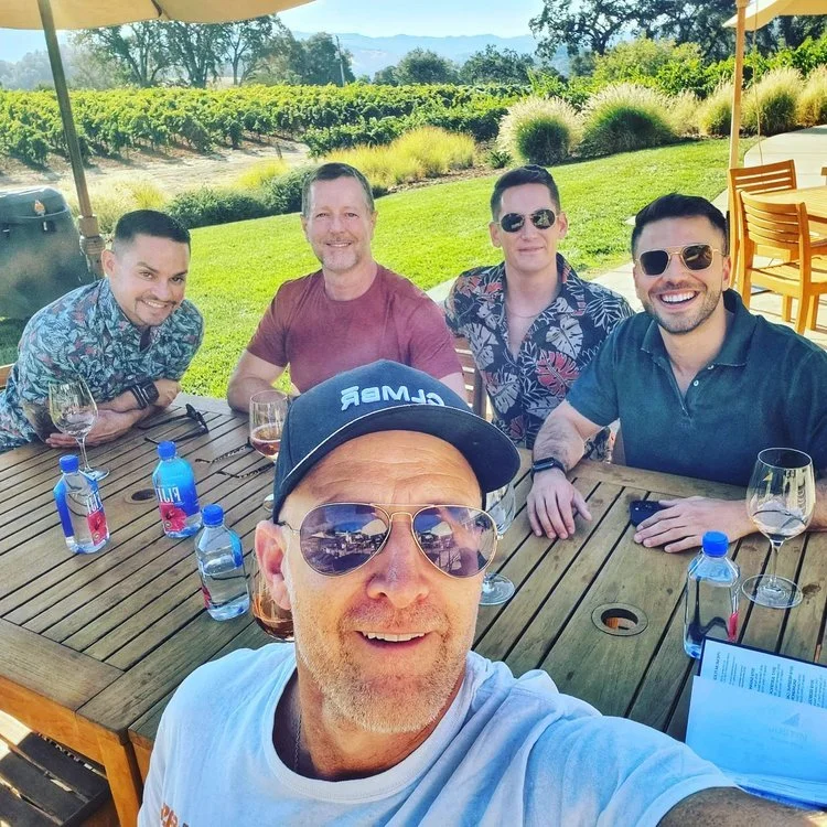 Five men sitting outdoors at a wooden patio table with wine glasses and water bottles, with a vineyard in the background under a sunny sky.