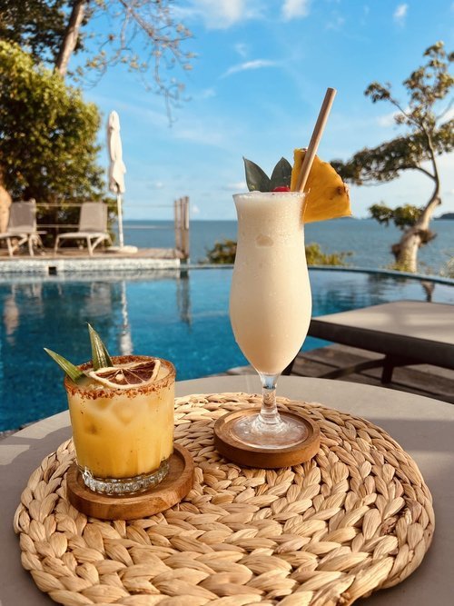 Two tropical cocktails on a wicker mat by a pool with ocean view, one garnished with pineapple and straw, the other with a dried citrus slice.