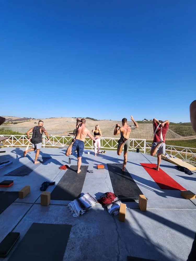Group of people practicing yoga on an outdoor terrace with mats and blocks, under a clear blue sky, in a rural landscape.